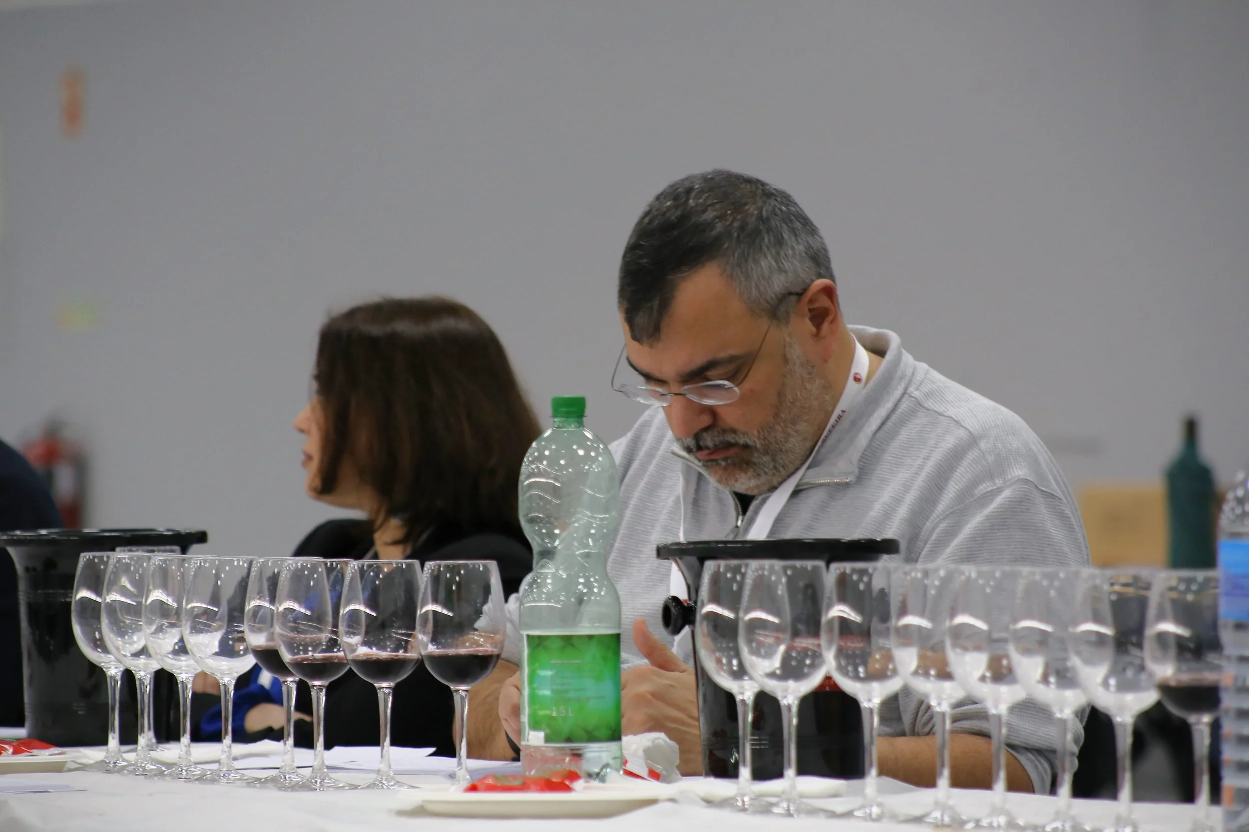 Homem com barba e óculos ajustando uma taça de vinho tinto em uma mesa com várias taças ao redor, em uma sala de eventos.