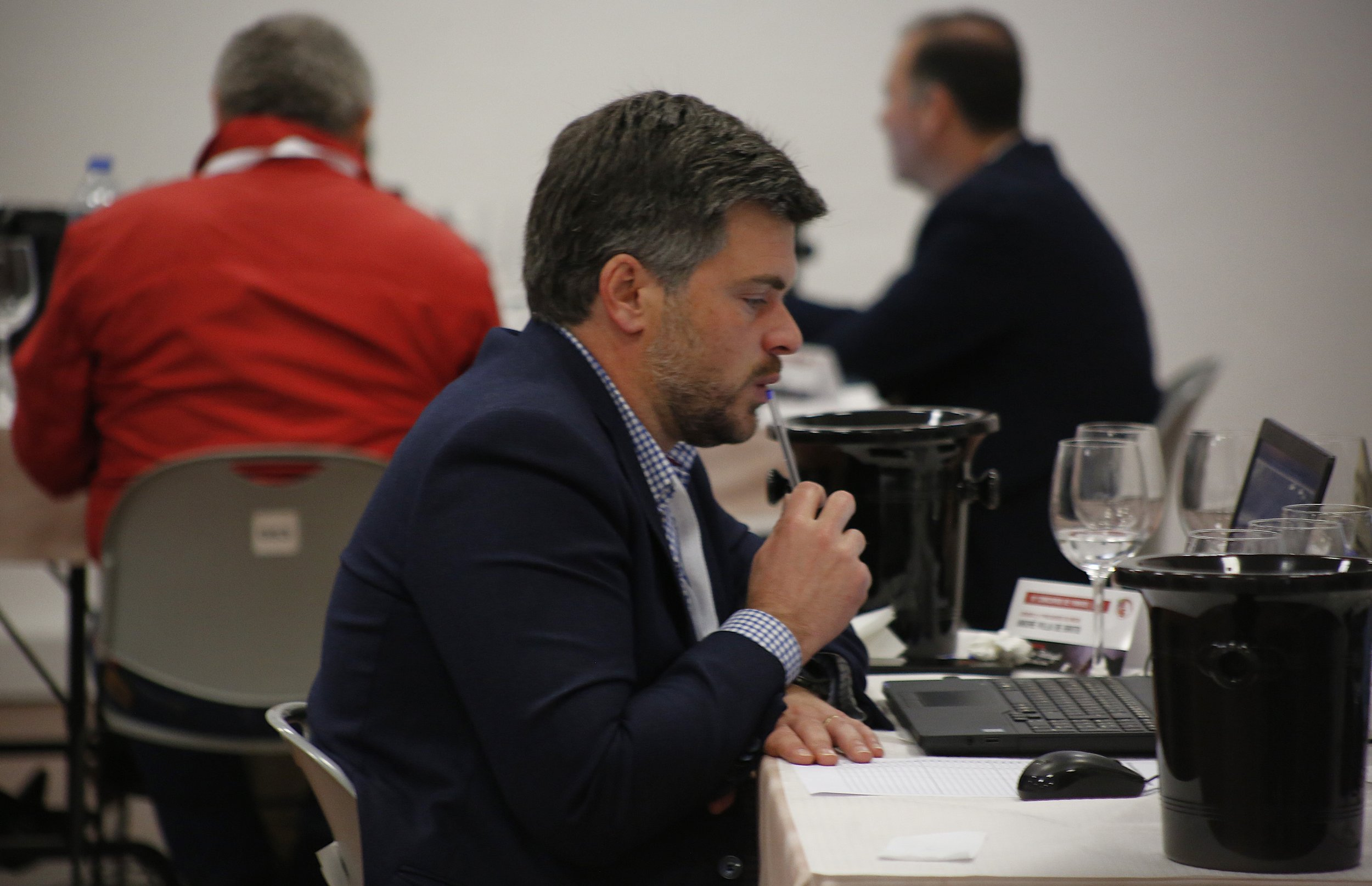 Homem de terno azul sentado na mesa de reunião, segurando uma caneta na boca, com laptops e copos de água na mesa. Ao fundo, duas outras pessoas também participando da reunião.