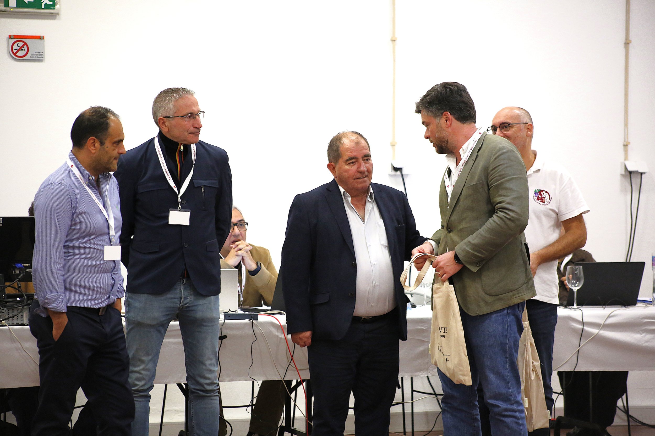 Homens em um encontro, alguns trocando presentes. Dois estão na frente, um entregando algo ao outro. Ao fundo, mais pessoas sentadas em uma mesa com laptops, copos e equipamentos eletrônicos.