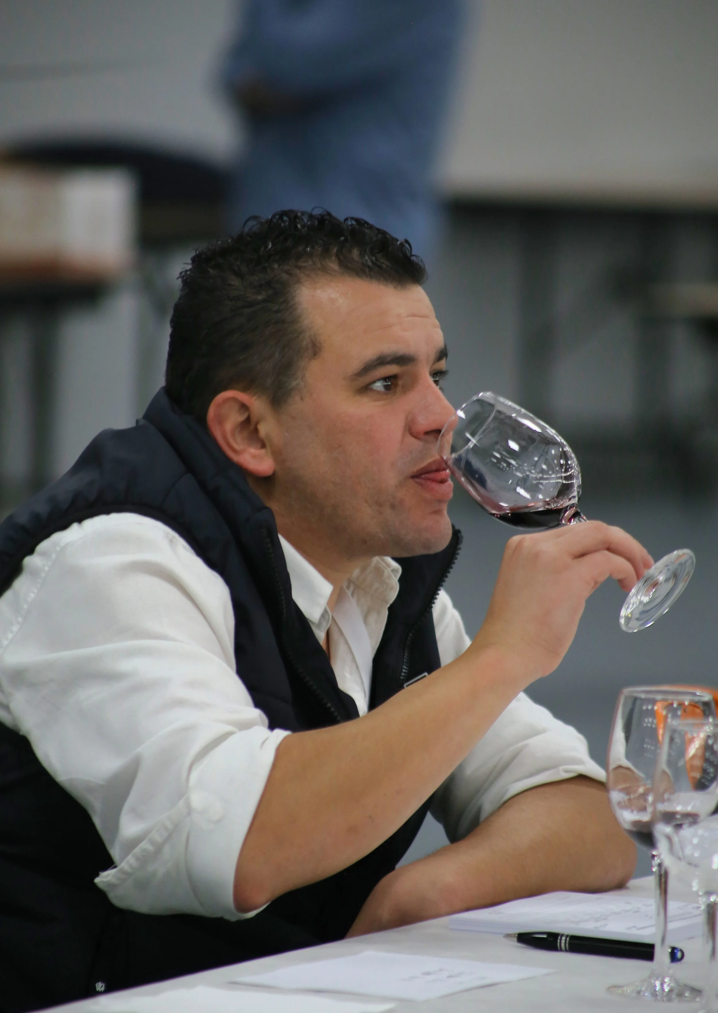 Homem de cabelo escuro e cabelo cacheado, vestindo camisa branca e colete preto, degustando vinho tinto em uma taça em uma mesa.