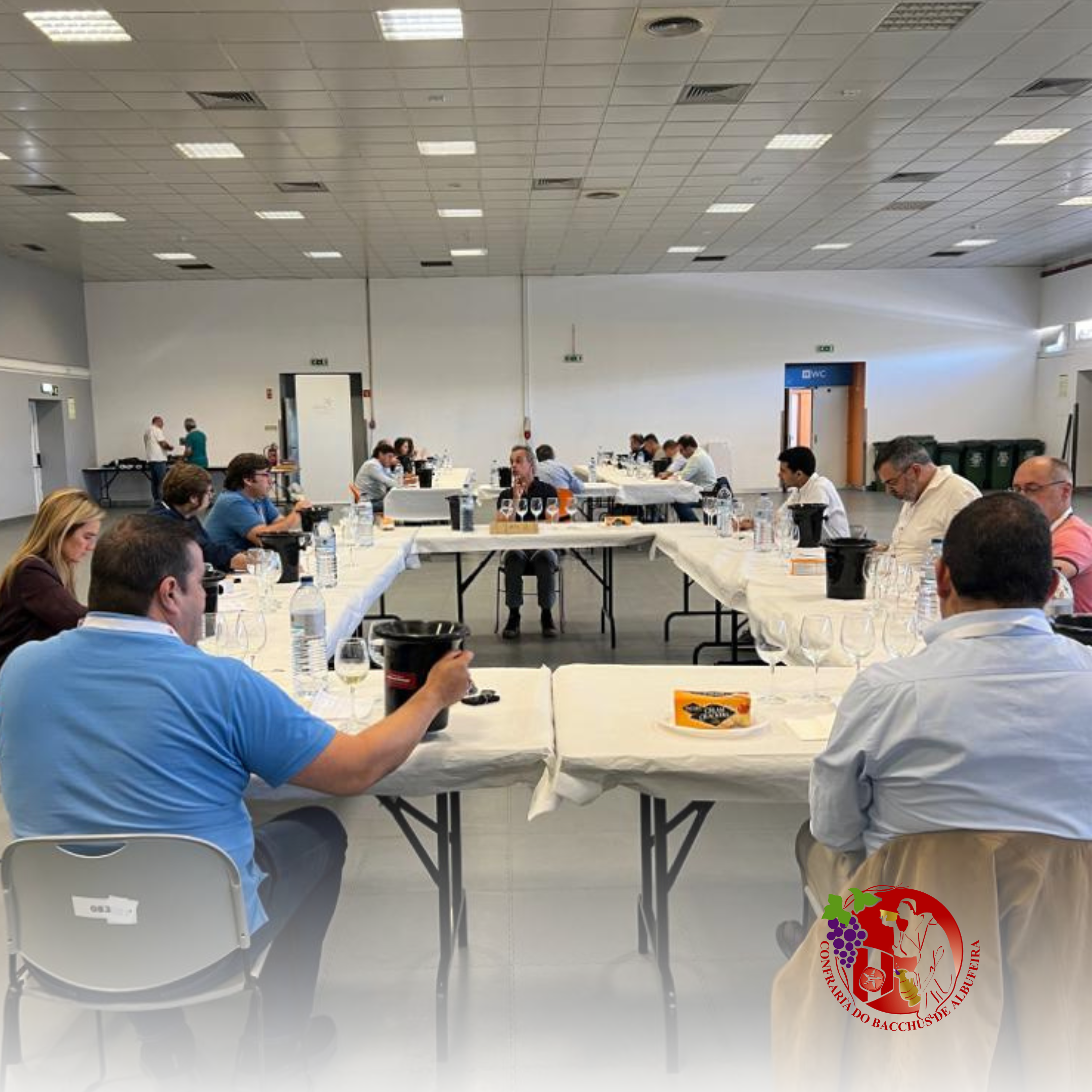 Reunião de pessoas ao redor de uma mesa longa, com copos de vinho, garrafas de água, e filtro de café, em uma sala de conferências, com uma porta azul ao fundo, e uma parede branca.