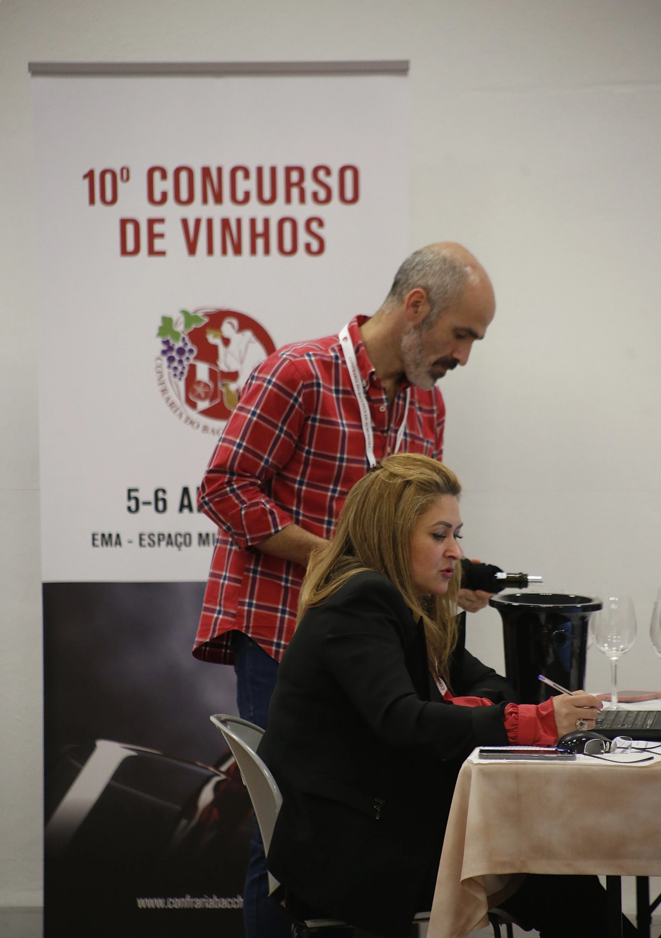 Homem e mulher participando de uma prova de vinhos em um evento com banner ao fundo.