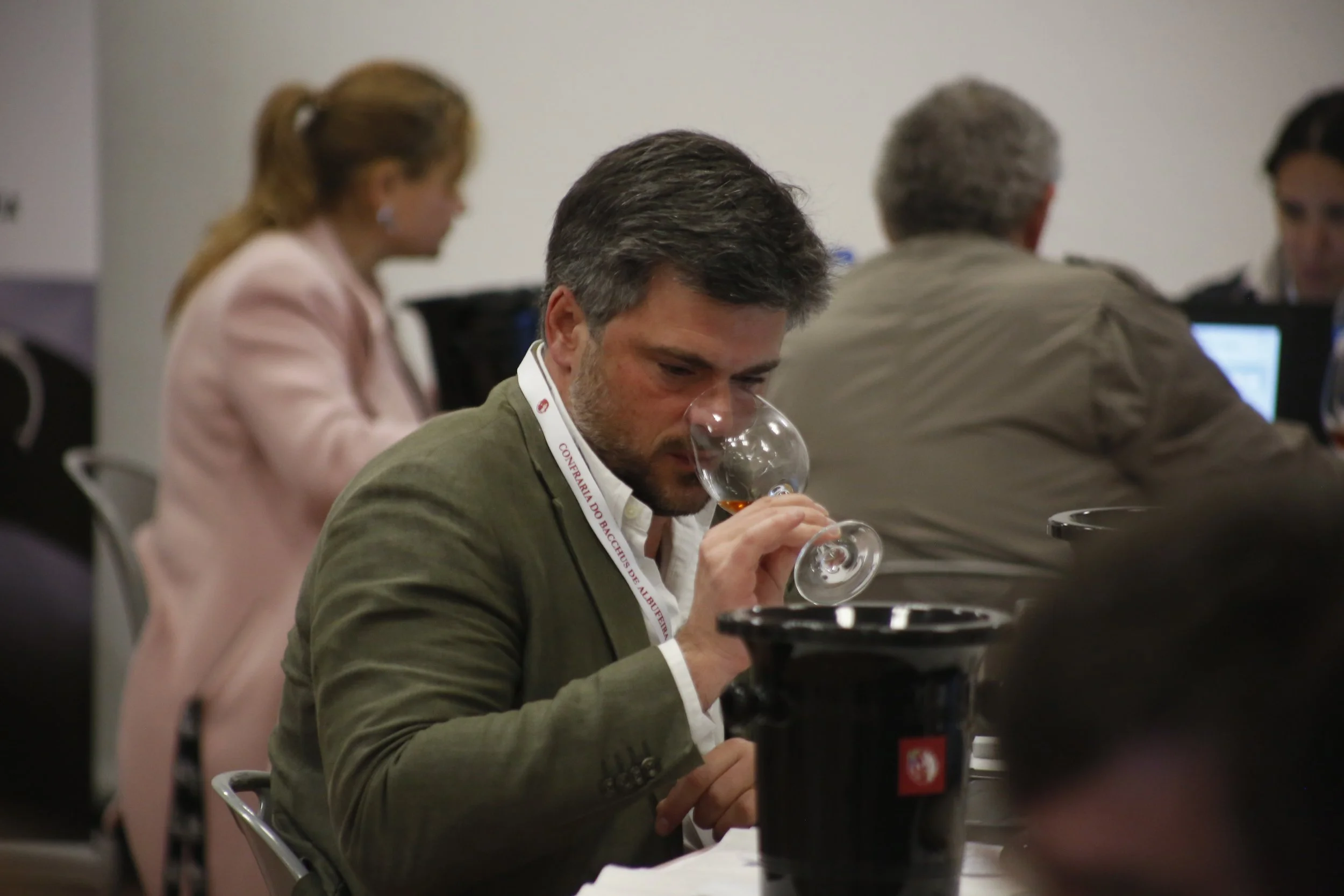 Homem provando uma taça de vinho, sentado em uma mesa em um evento de degustação ou conferência, com outras pessoas ao fundo usando computadores.