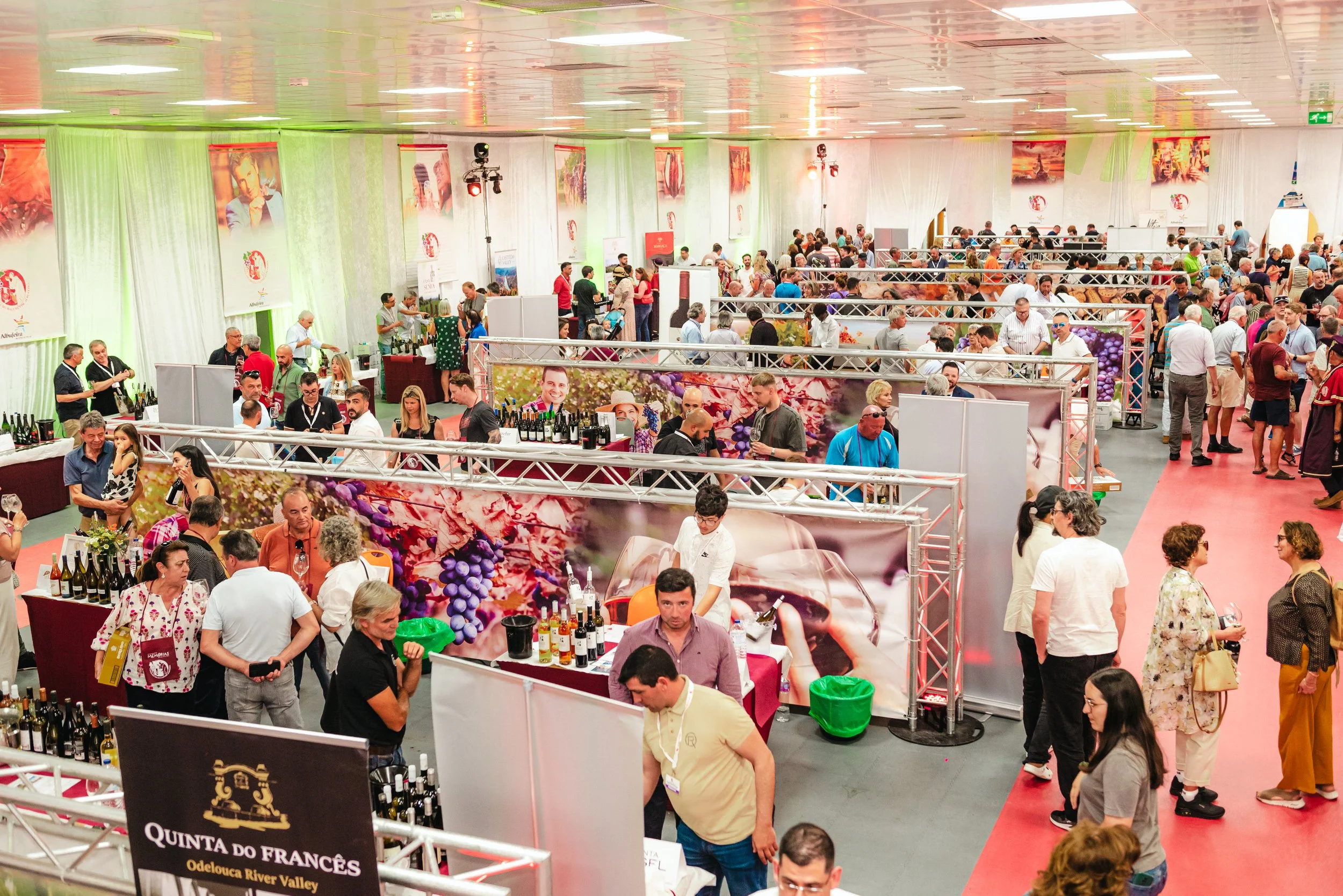 Grande feira de vinhos com várias barracas e visitantes em um espaço coberto. Pessoas degustando e conversando, banners e iluminação decorativa.