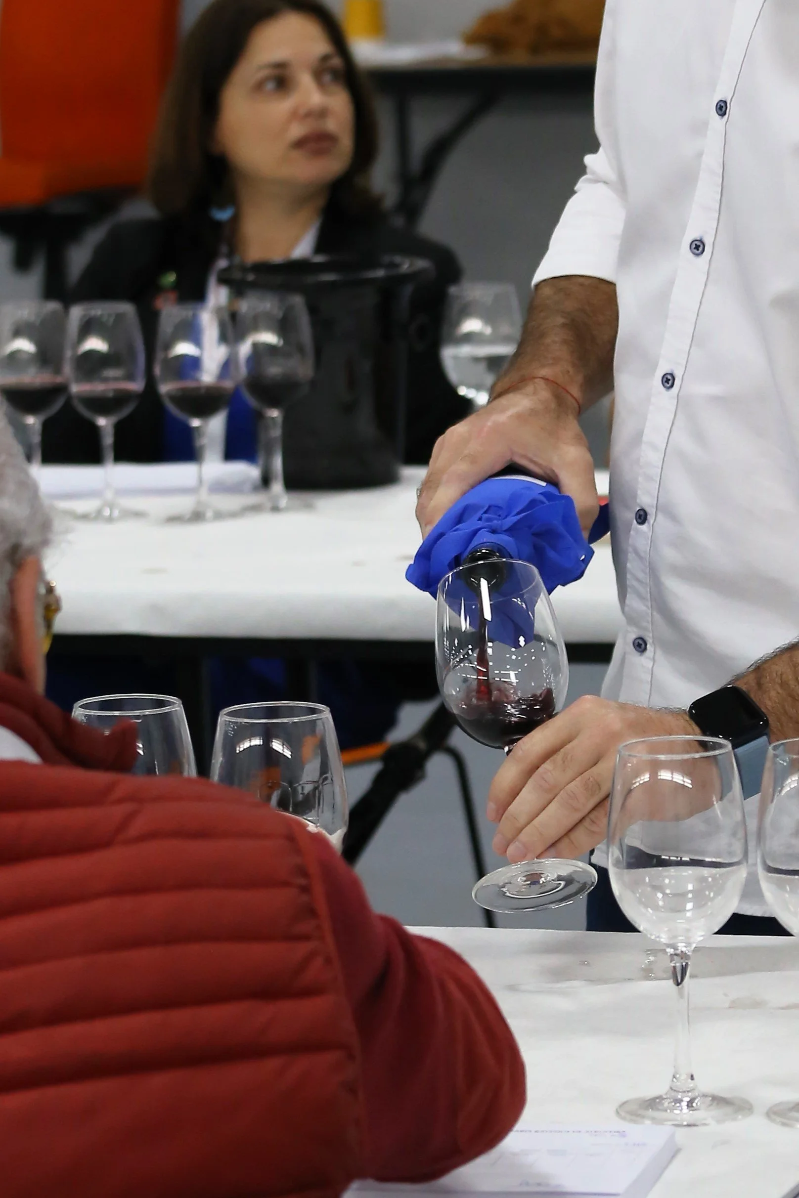 Pessoa despejando vinho tinto em uma taça durante uma degustação, com várias taças de vinho na mesa e outras pessoas ao redor.