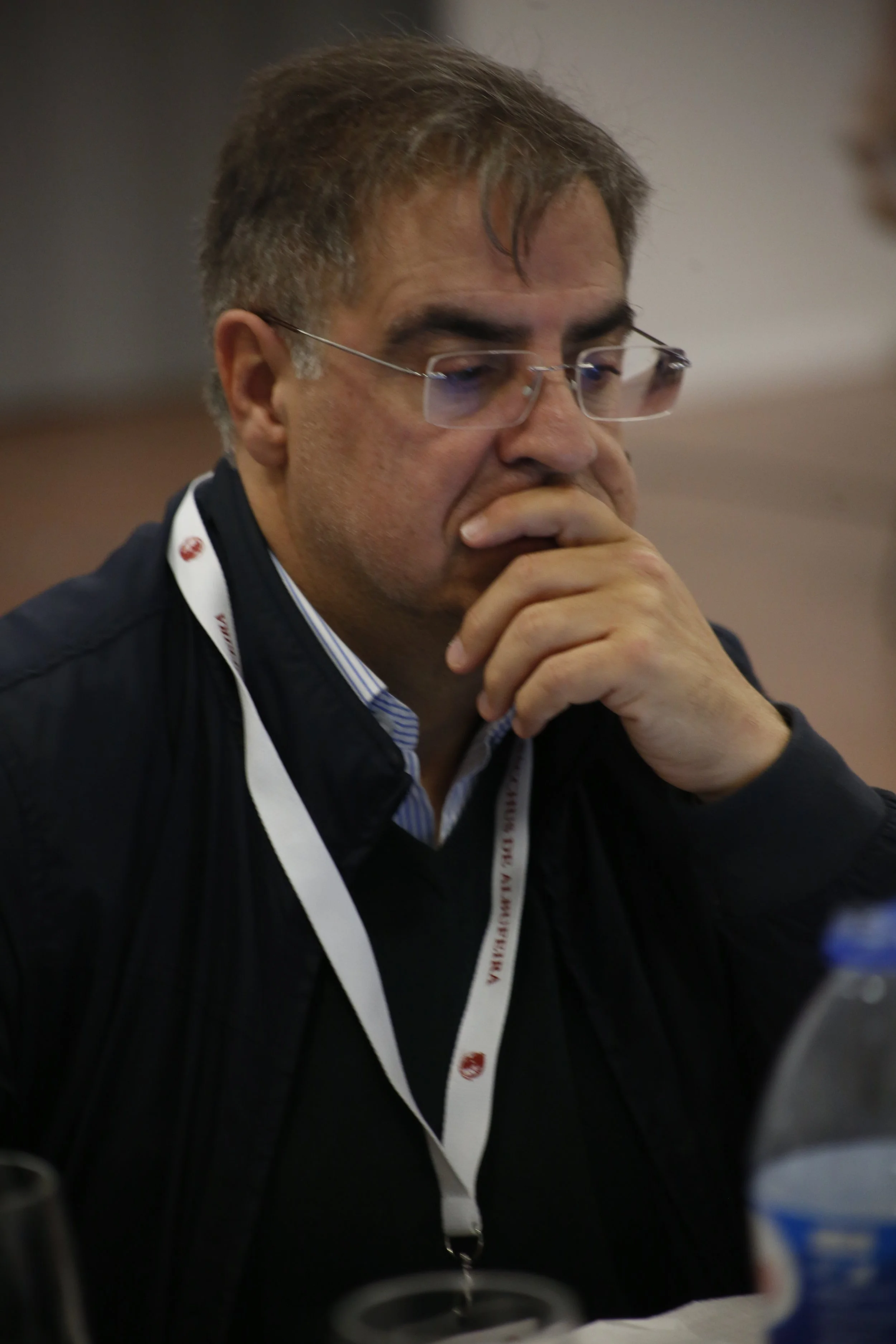 Homem pensativo com óculos, vestindo jaqueta escura e camiseta de colarinho, sentado em ambiente interno com foco em um evento ou reunião, usando crachá ao redor do pescoço.