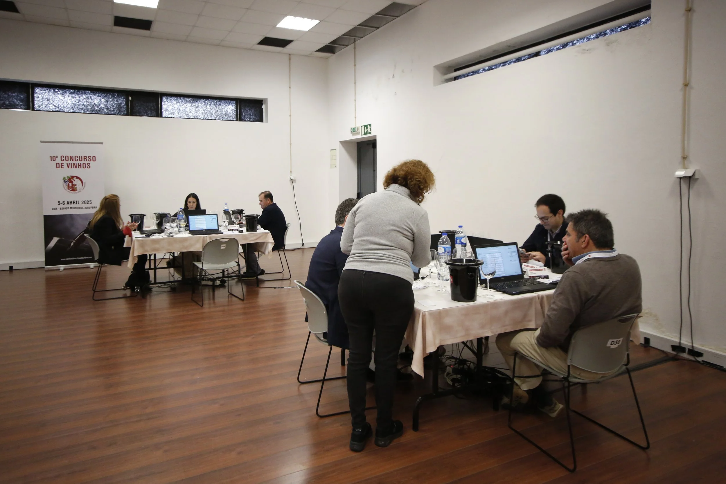 Pessoa conversando com um grupo de pessoas em uma sala, com uma mesa de reunião, computadores e garrafas de água.