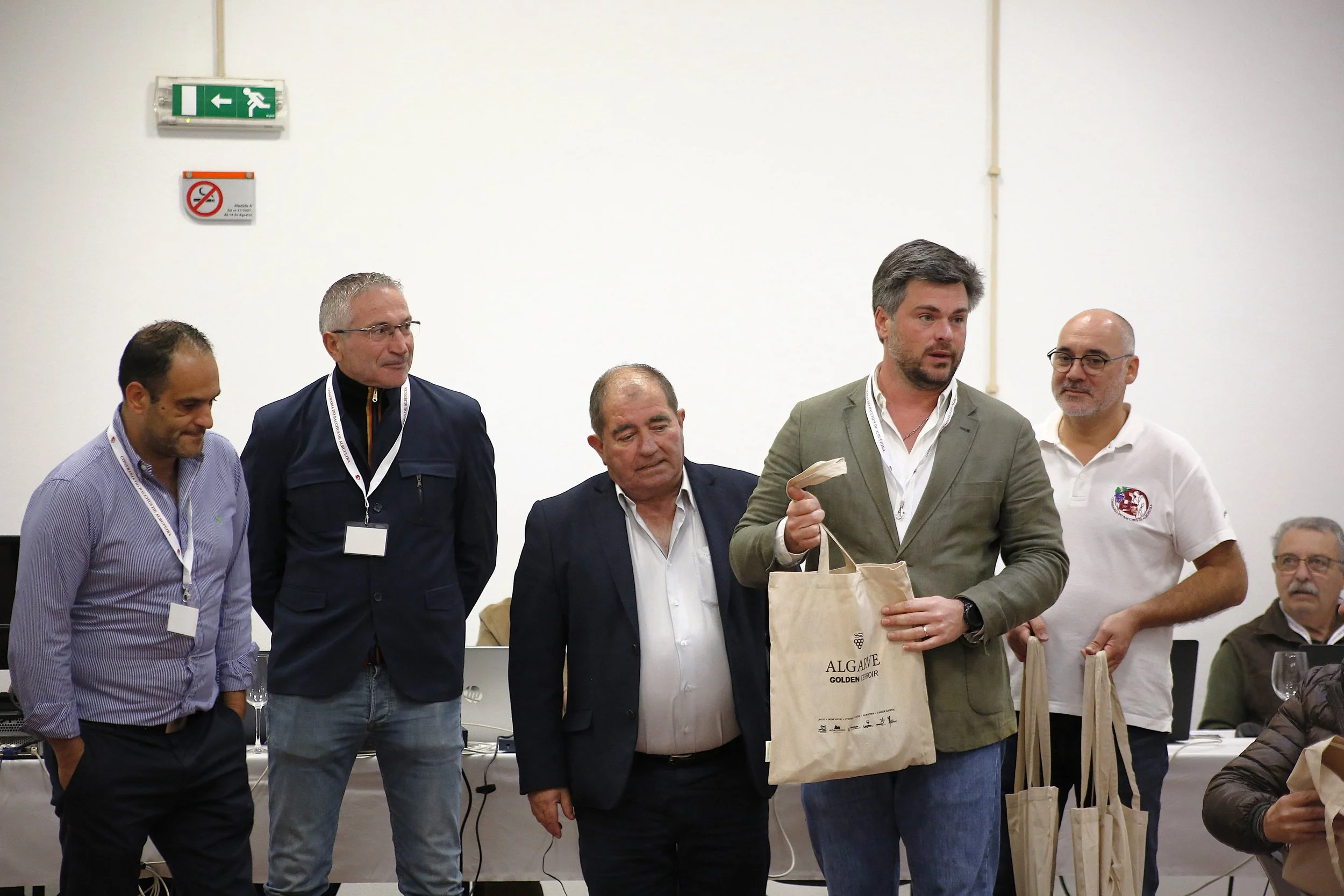 Grupo de homens participando de uma cerimônia, alguns segurando sacolas de presente, em um ambiente interior com fundo branco e uma mesa ao fundo.