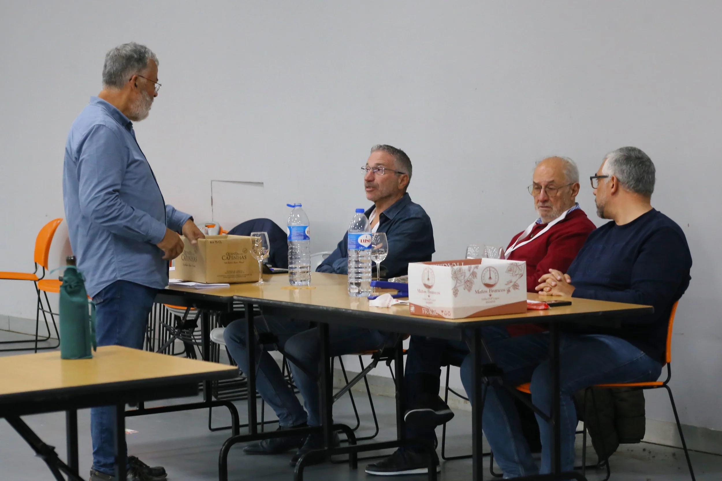 Quatro homens com cabelos grisalhos estão sentados à mesa em uma sala branca, enquanto um homem com cabelo grisalho e barba, de óculos, está de pé ao lado da mesa, manuseando uma caixa de papelão.