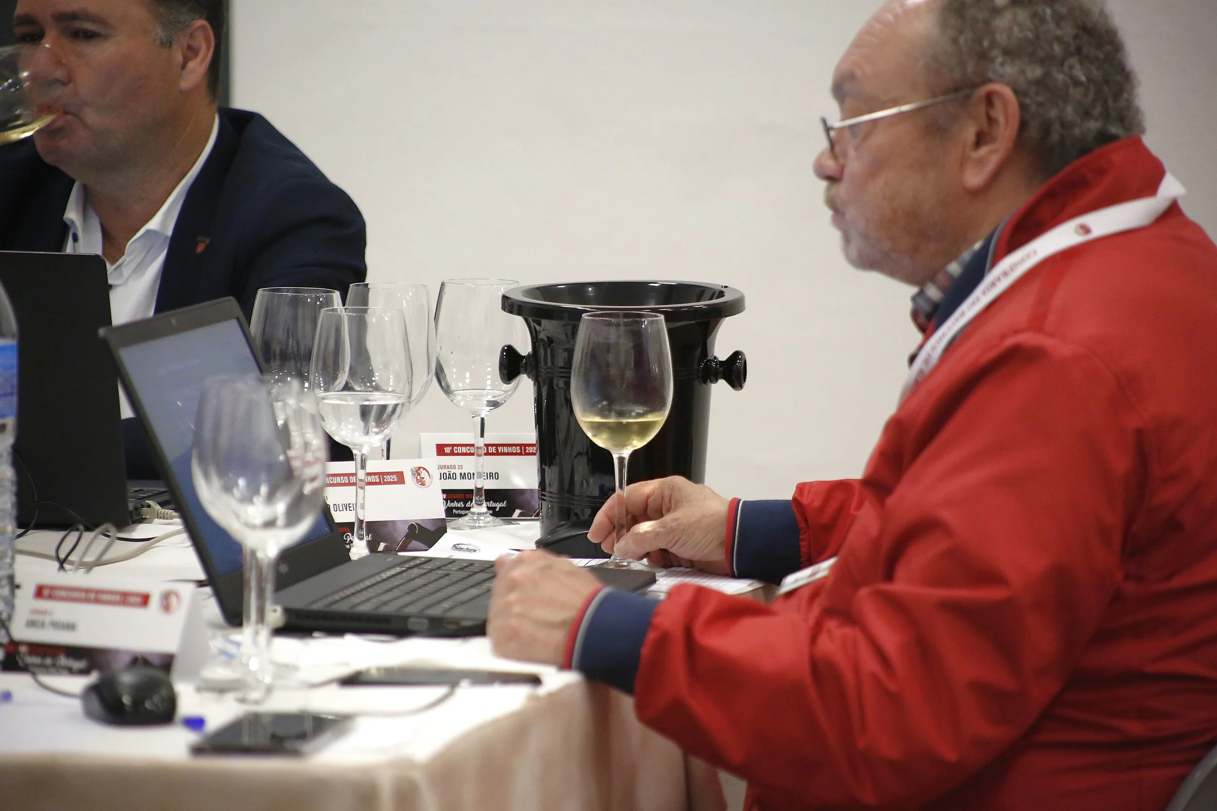Homens participando de degustação de vinhos, um deles segurando uma taça com vinho branco, em uma mesa com várias taças, laptops, um balde para vinho, e apontador de cartas de classificação de vinhos.