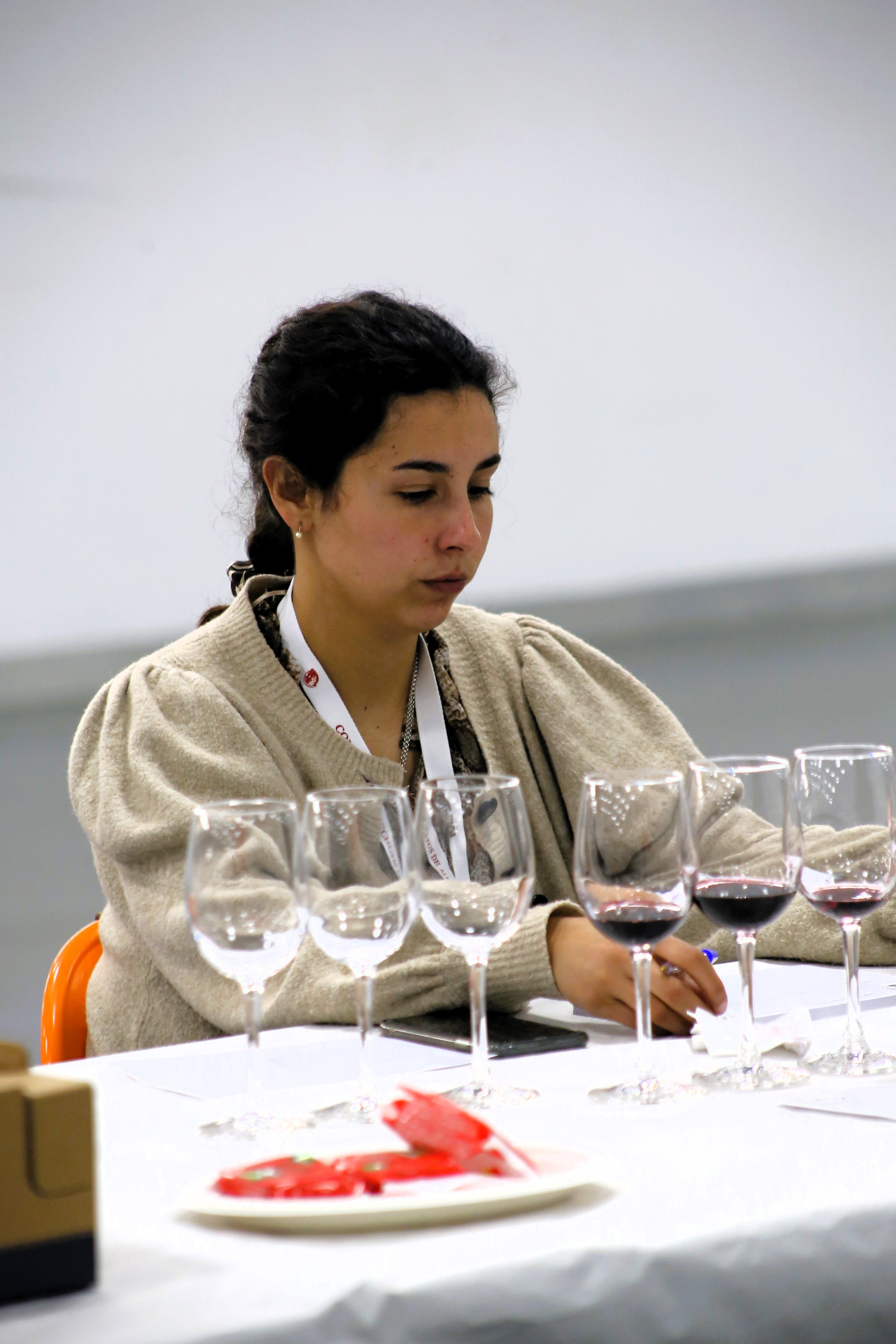 Mulher sentada em uma mesa com taças de vinho. Ela parece estar em uma degustação de vinhos, observando atentamente suas taças.