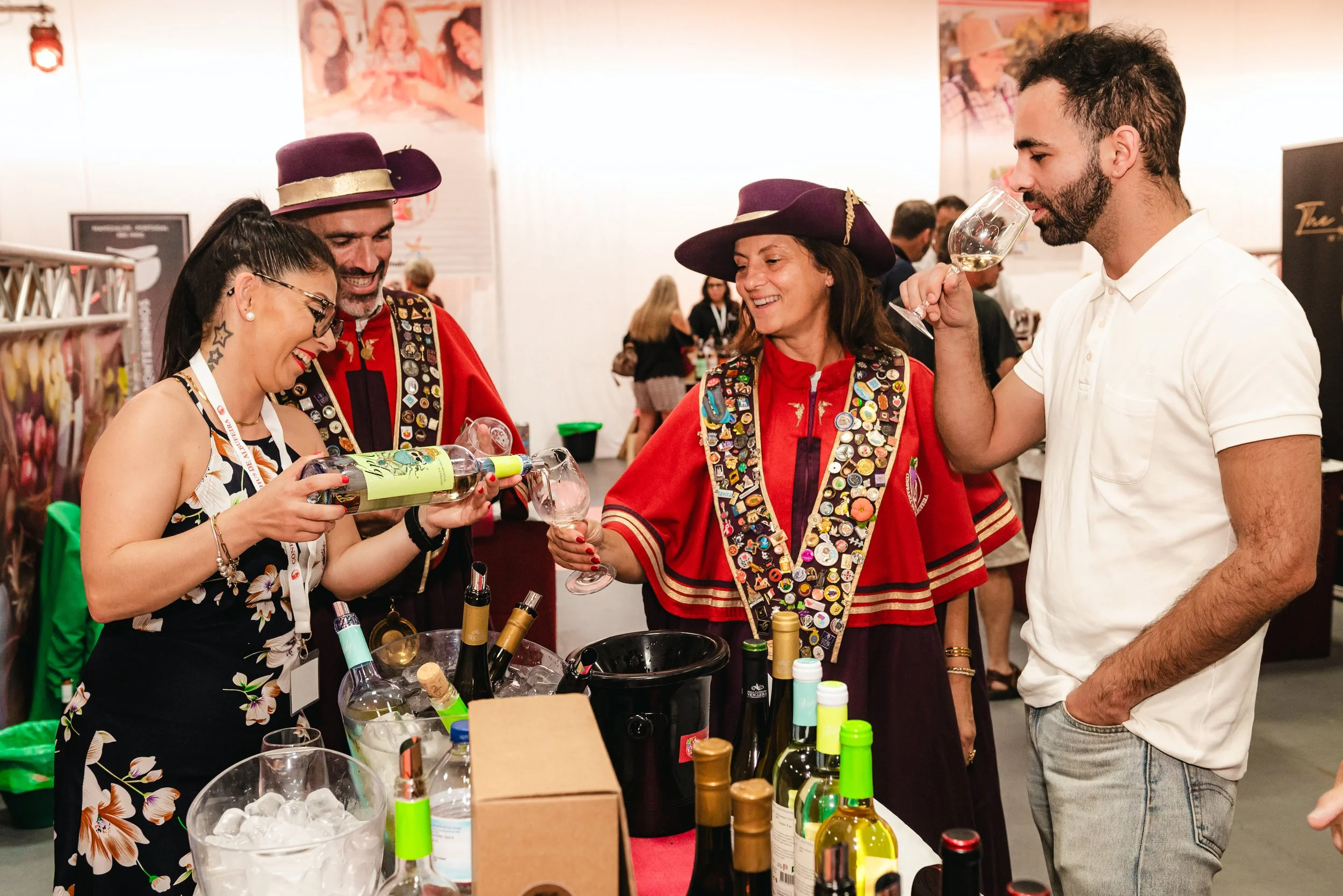 Pessoas em uma feira de vinhos, duas vestidas com roupas tradicionais de escoteiro, uma mulher em traje com muitas bottons, uma jovem com óculos escuros e um homem em camiseta branca, todos segurando taças de vinho.