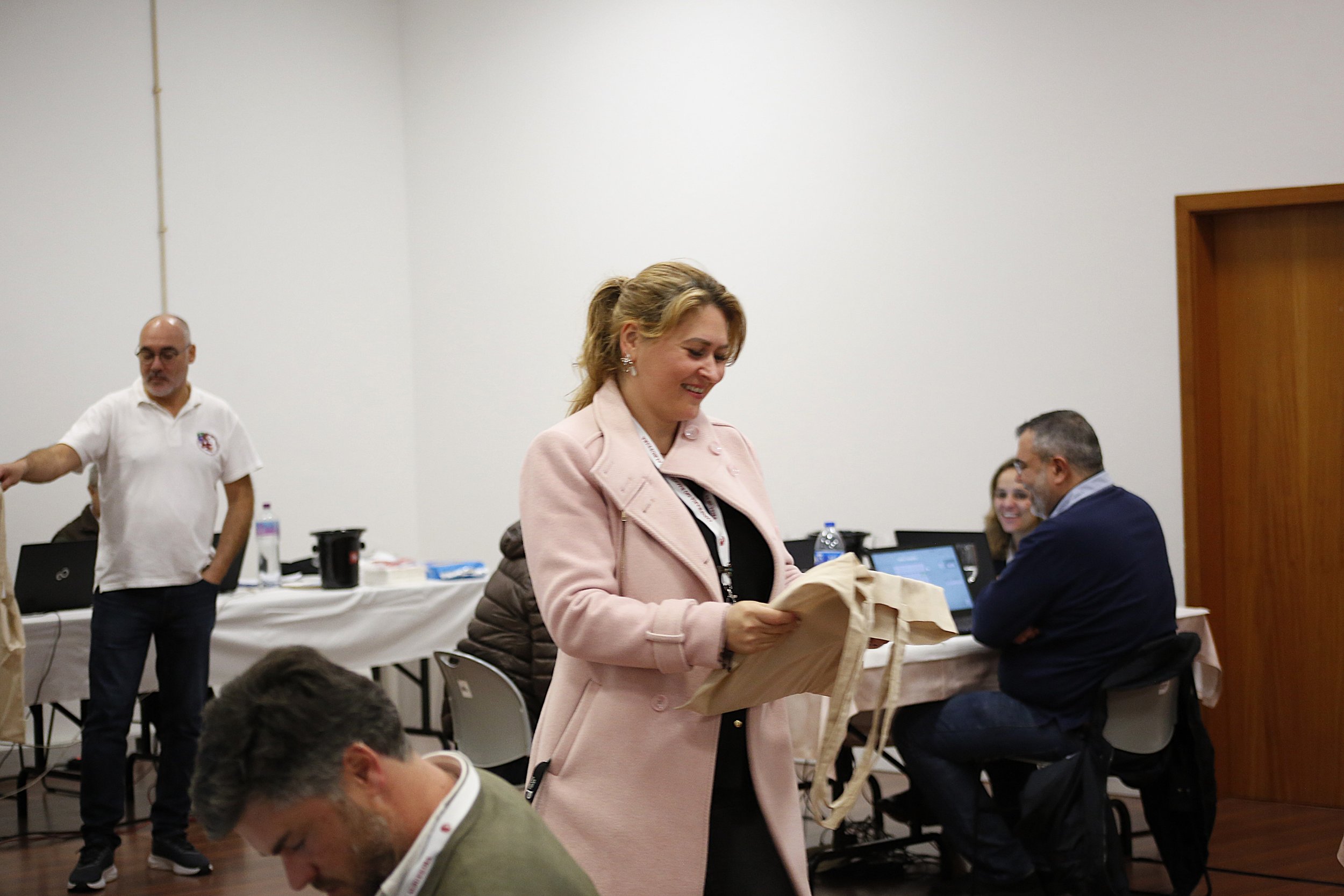 Mulher sorridente de cabelos ondulados, vestindo jaqueta rosa claro, segurando uma sacola de papel, em ambiente de reunião com várias pessoas ao redor, computadores e materiais na mesa.