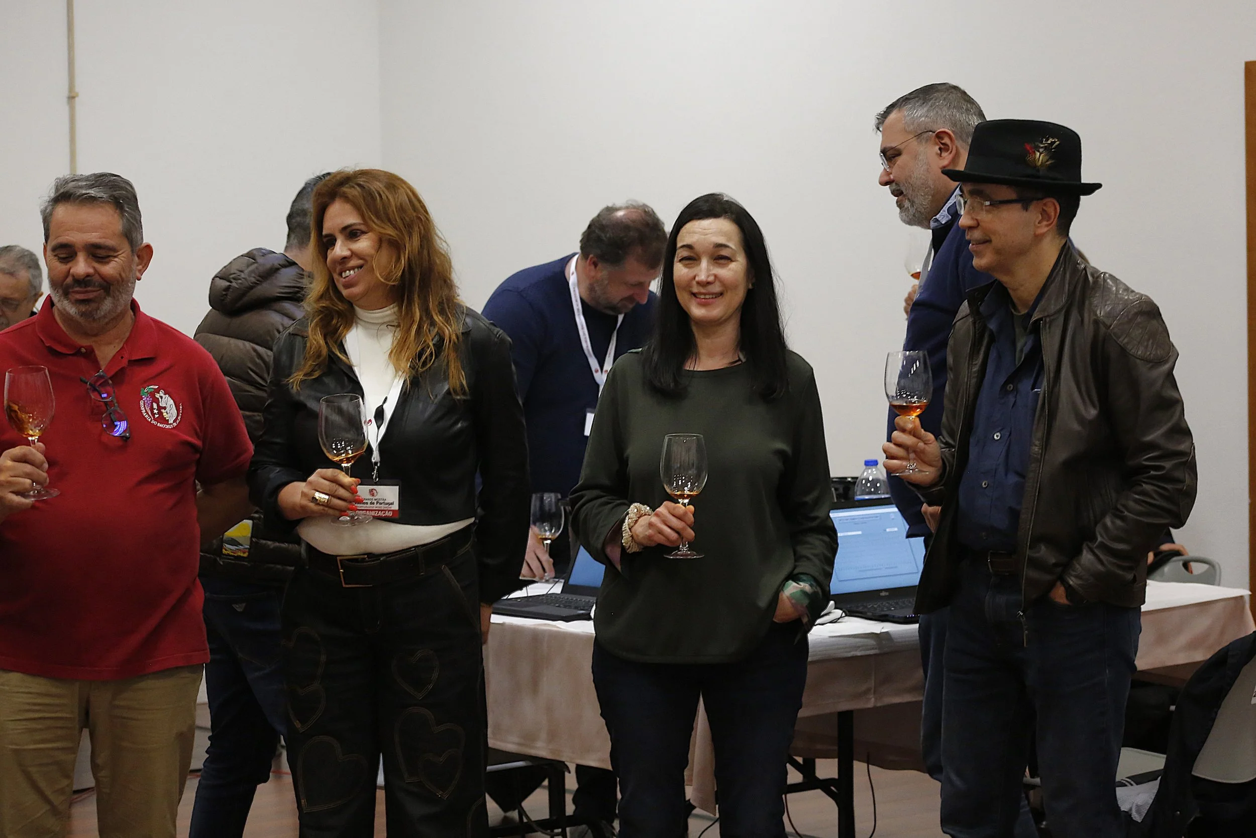 Grupo de pessoas sorrindo e conversando enquanto seguram taças de vinho em uma festa ou evento, com algumas pessoas ao fundo ao redor de uma mesa com computadores.