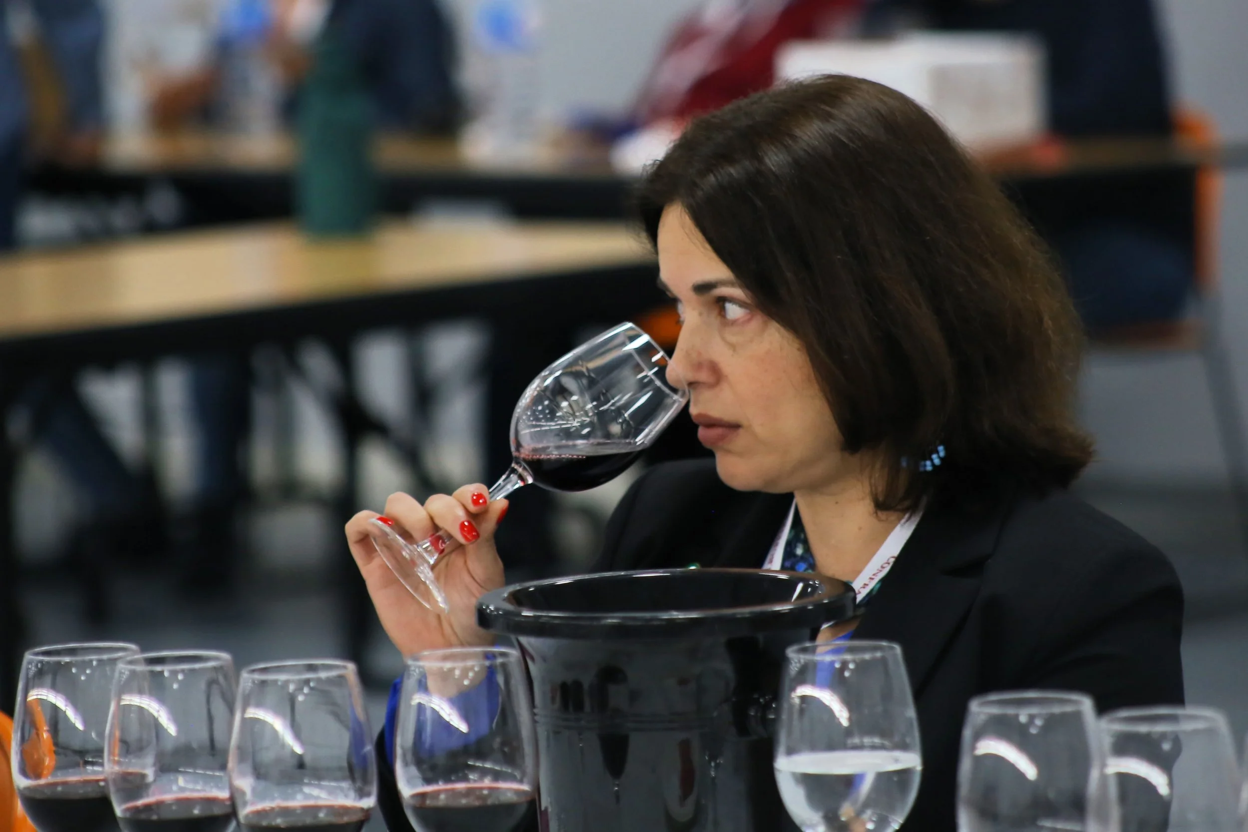 Mulher com cabelo castanho escuro segurando uma taça de vinho tinto, cheirando o vinho, em uma mesa com várias taças de vinho e um balde de gelo ao seu redor em uma sala de evento.
