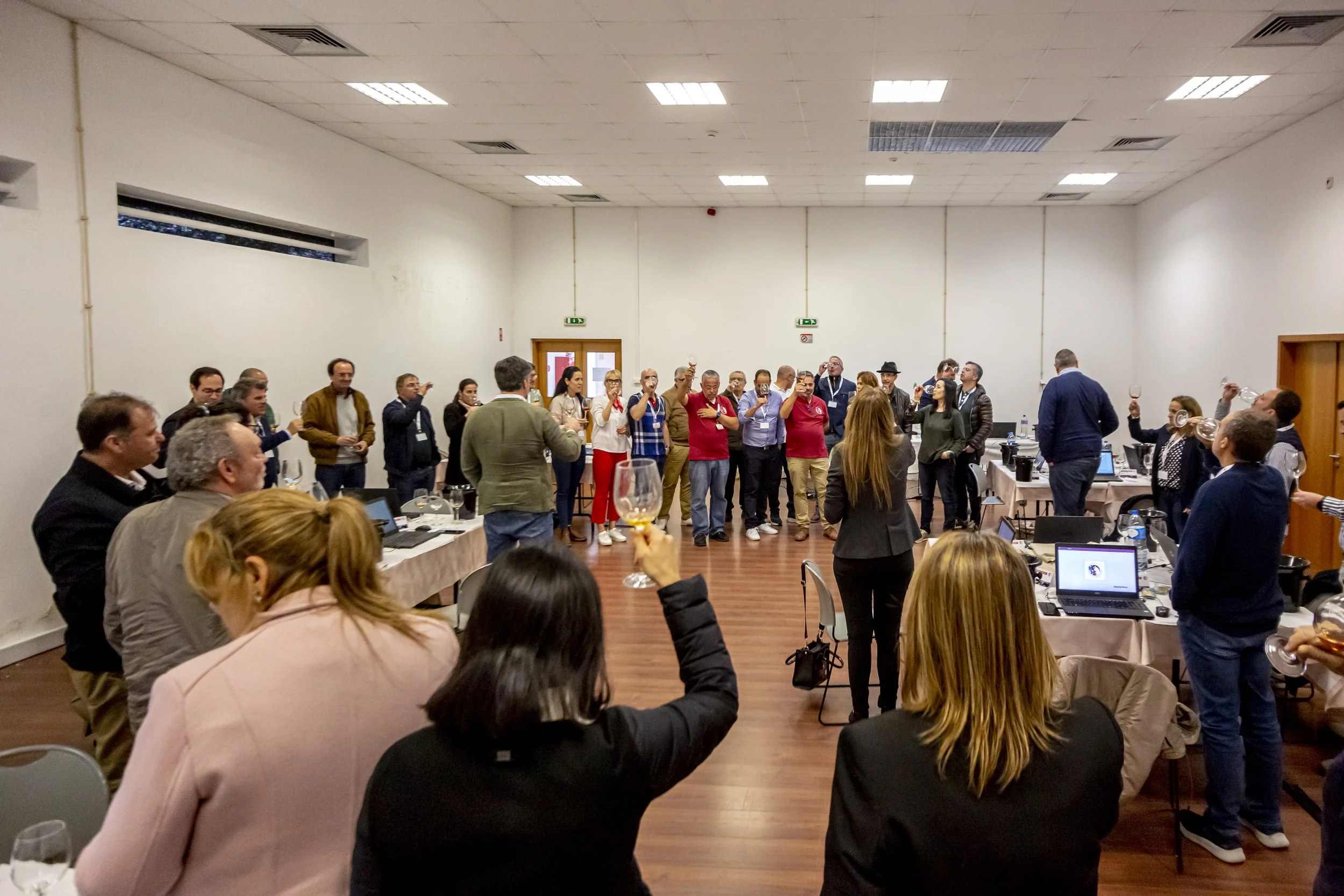 Grupo de pessoas em evento de comemoração em uma sala grande, muitas delas levantando taças de vinho, algumas sentadas e outras em pé, com um ambiente de reunião ou confraternização.