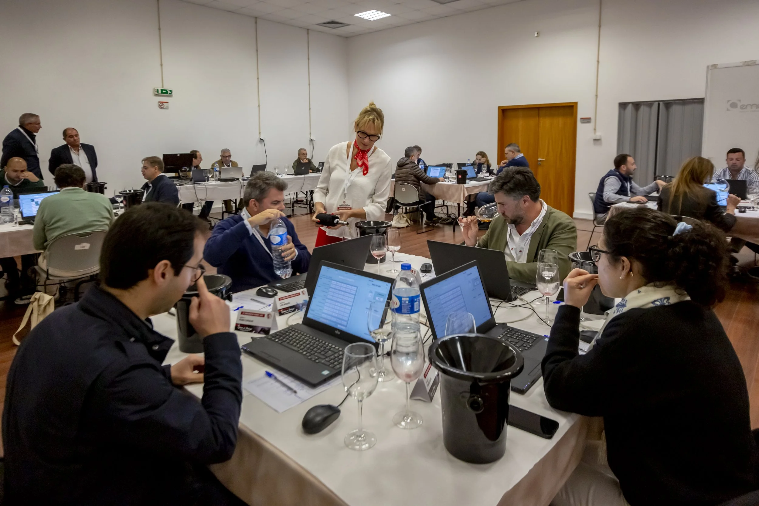 Reunião de negócios com várias pessoas usando laptops, em uma sala de conferências, com uma mulher de óculos e cabelo loiro no centro, interagindo com os participantes.