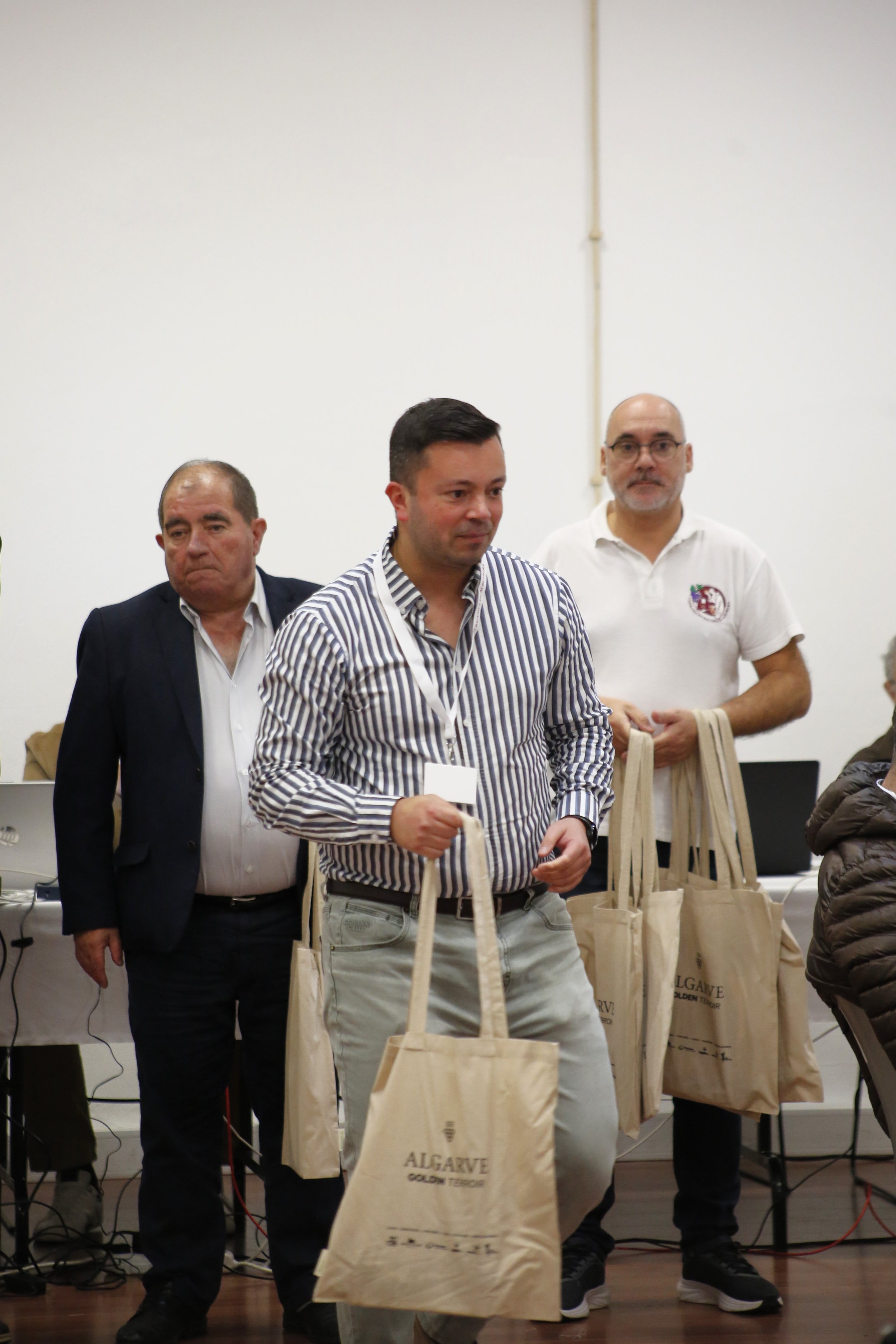 Homem recebendo sacolas de papel na frente de uma parede branca, com outros homens ao fundo, em um evento indoor.