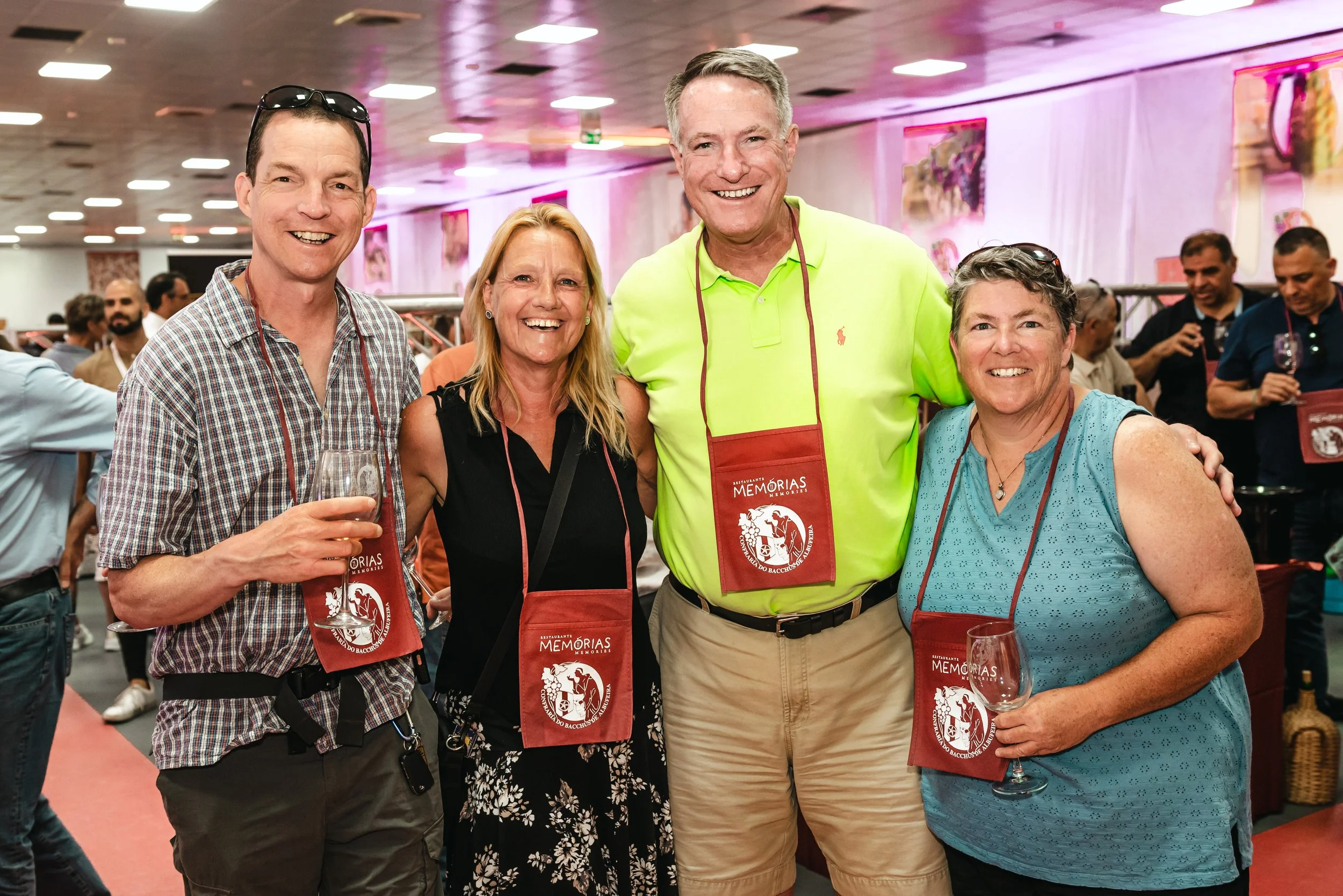 Grupo de pessoas sorrindo em evento social, usando aventais vermelhos com o logotipo de memórias, ambiente com decoração rosa e várias pessoas ao fundo.