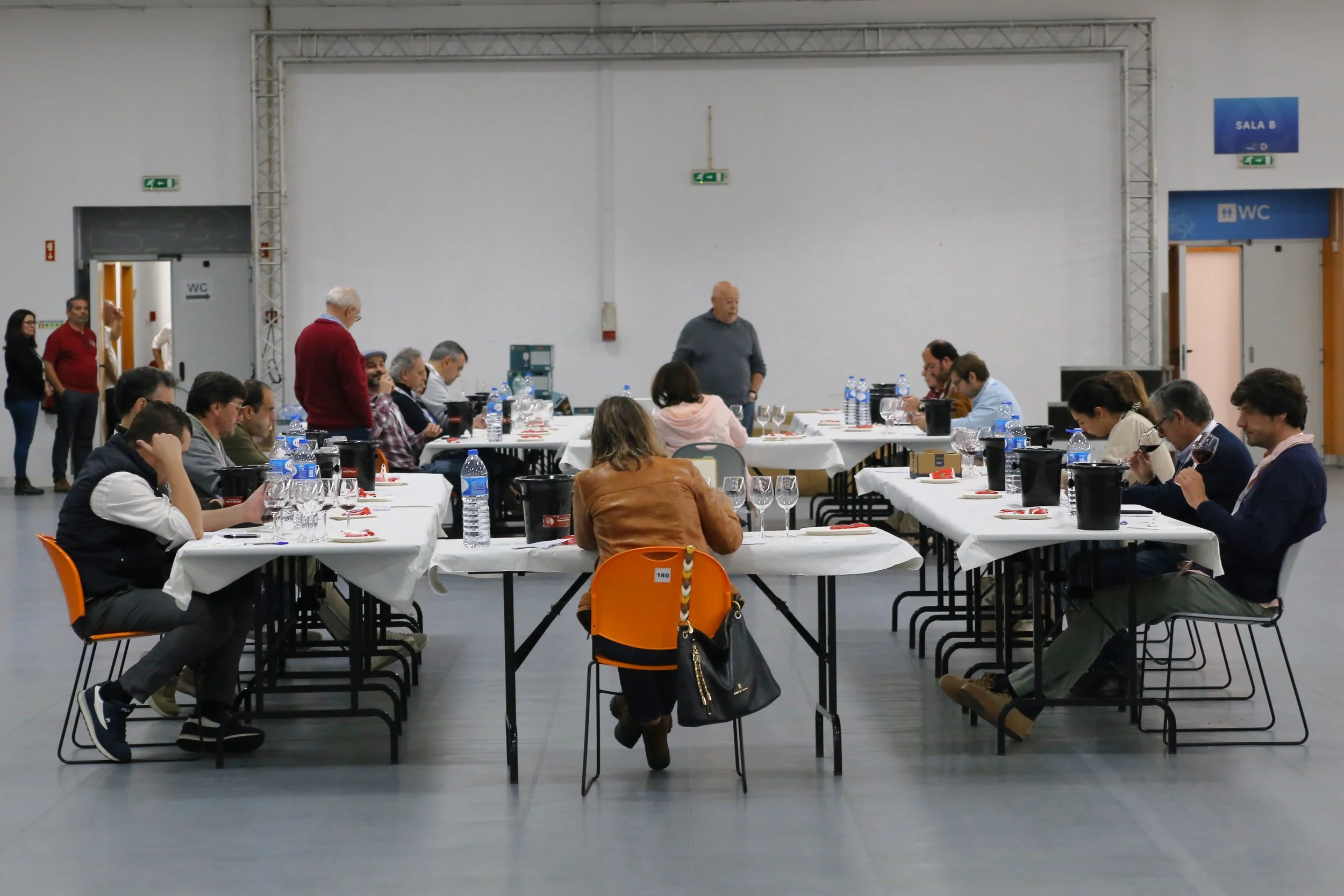 Pessoas sentadas em uma mesa de degustação de vinhos em uma sala ampla, com duas portas de banheiros ao fundo, uma das portas com sinalização de WC e outra com sinal de sala B.
