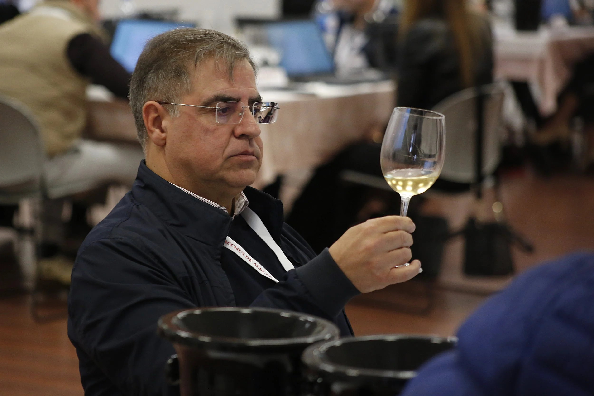 Homem segurando uma taça de vinho branco, com expressão séria, em ambiente de evento, com computadores e pessoas ao fundo.