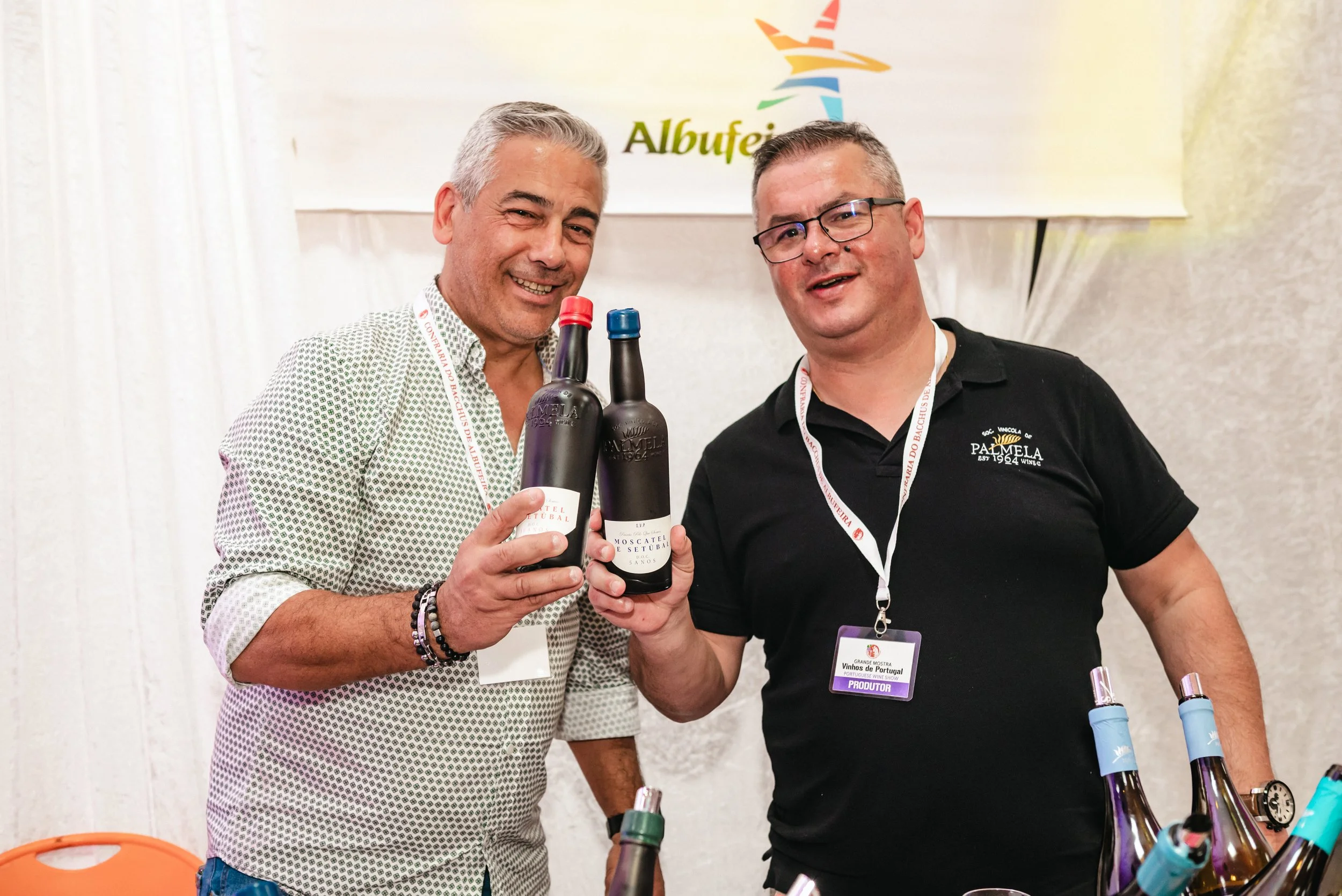 Dois homens segurando garrafas de vinho em uma feira, com nomes de produtores e logotipo de evento ao fundo.