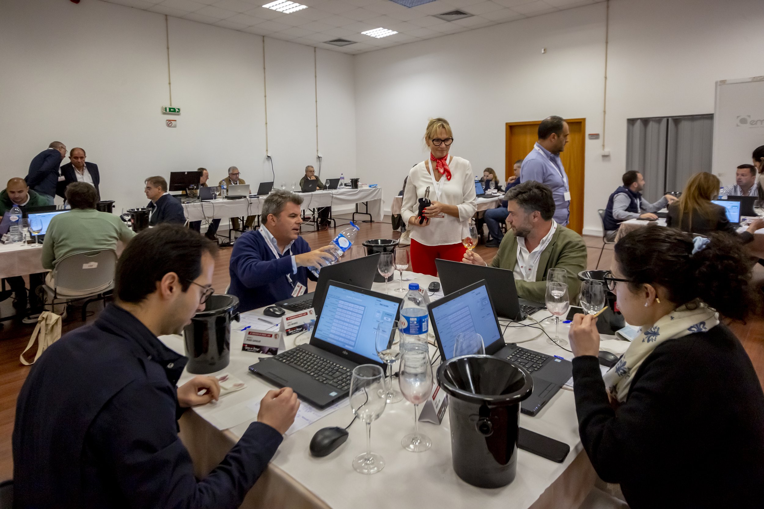 Pessoa apresentando-se a um grupo de pessoas em uma conferência ou reunião de trabalho, com várias pessoas usando laptops e dispositivos eletrônicos em uma sala de reunião.