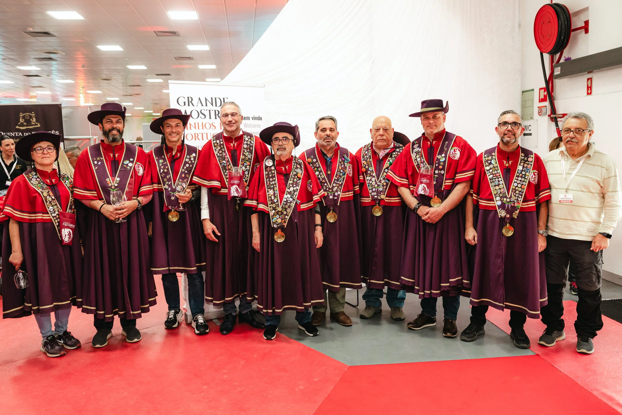 Grupo de pessoas fantasiadas com roupas tradicionais de gala, usando chapéus pretos, colares de medalhas e roupas vermelhas com detalhes dourados, posando juntas em um evento. Algumas pessoas seguram taças e todas estão na frente de uma parede branca