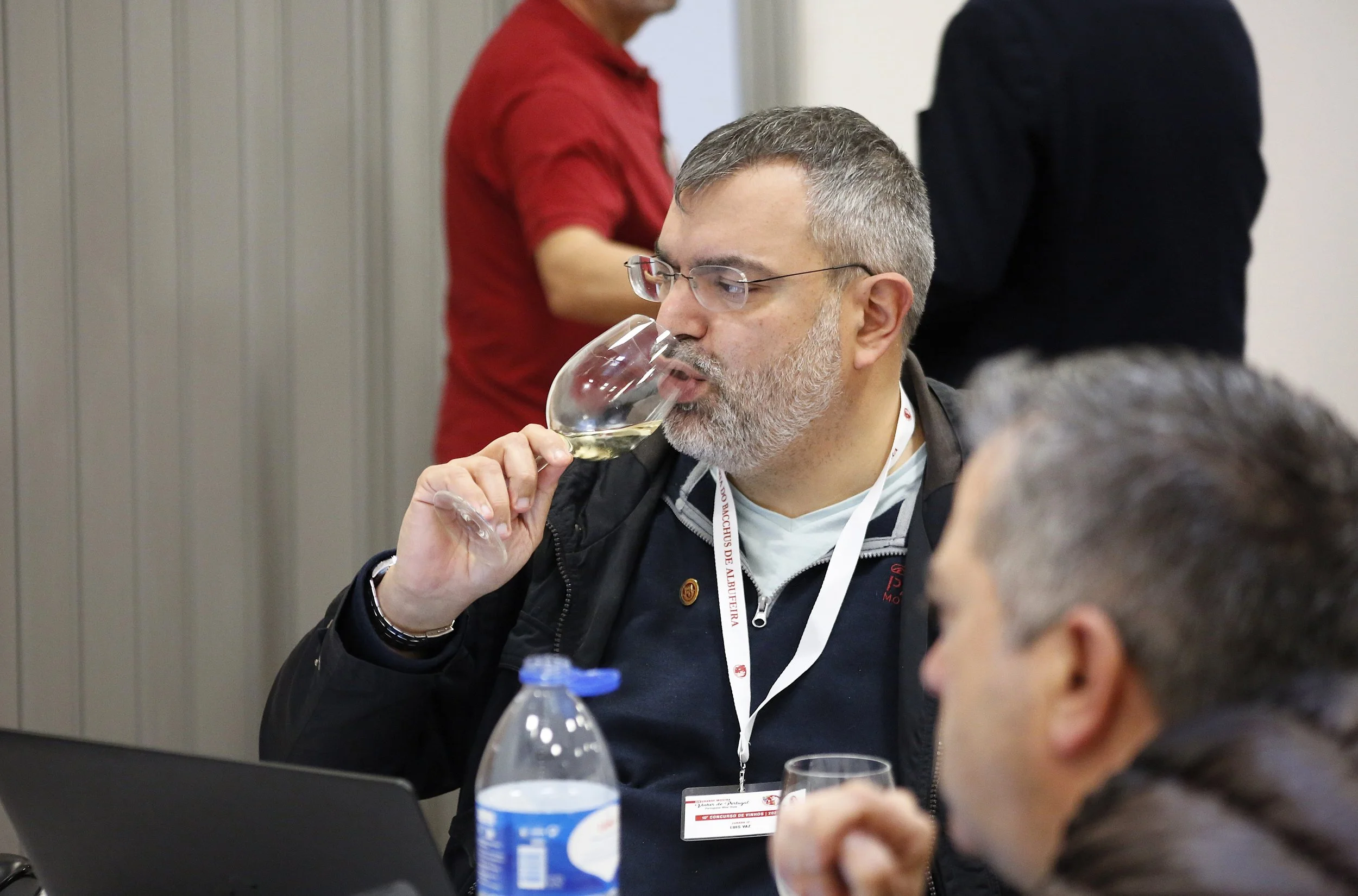 Homem com barba e óculos, usando uma jaqueta preta, está provando vinho branco em uma taça, sentado a uma mesa com uma garrafa de água e um laptop. Ao fundo, duas pessoas estão de pé, uma com camisa vermelha e outra com casaco preto.