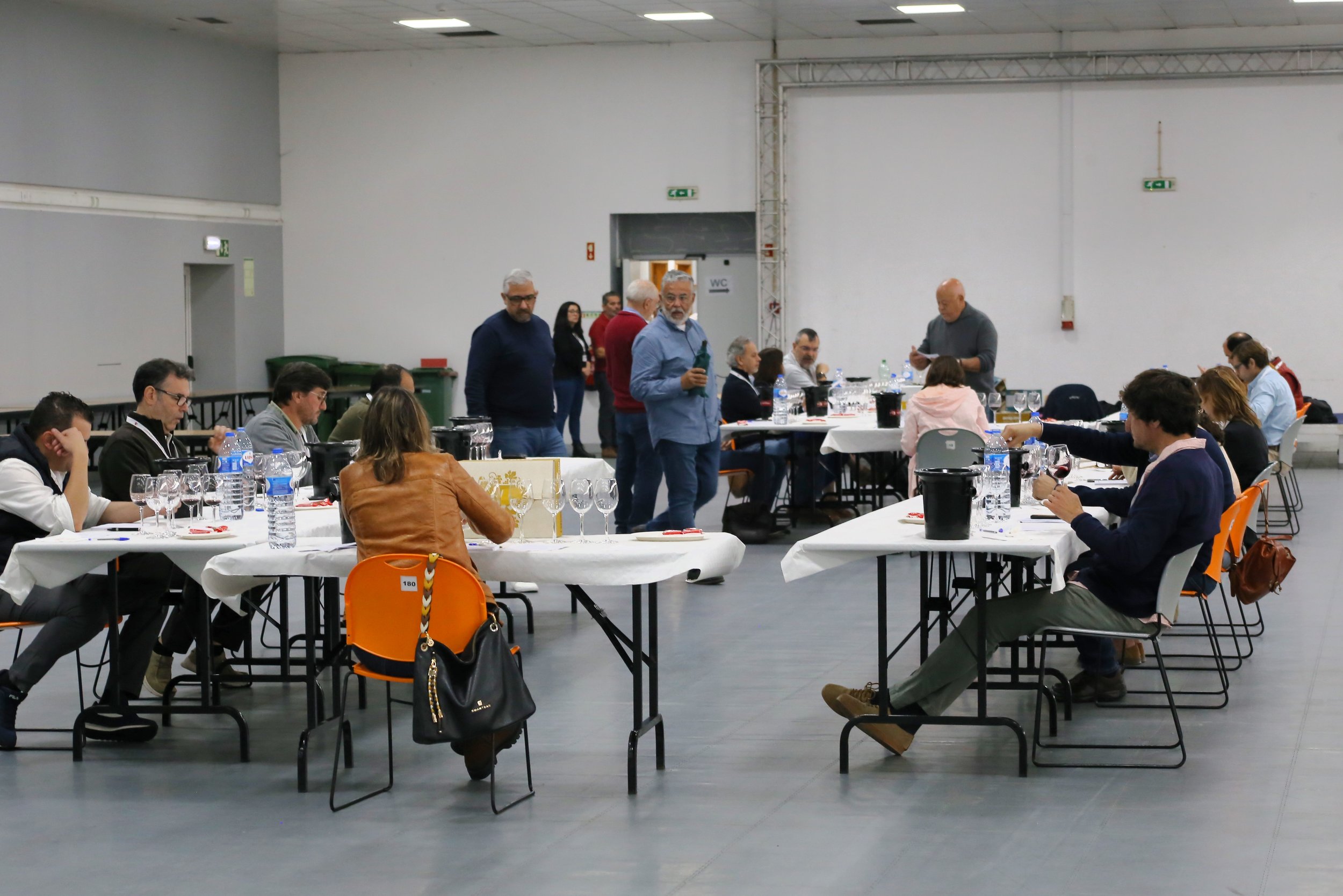 Reunião de pessoas em uma sala grande, sentadas ao redor de mesas com copos de vinho, garrafas de água e pratos, durante uma confraternização ou evento social.