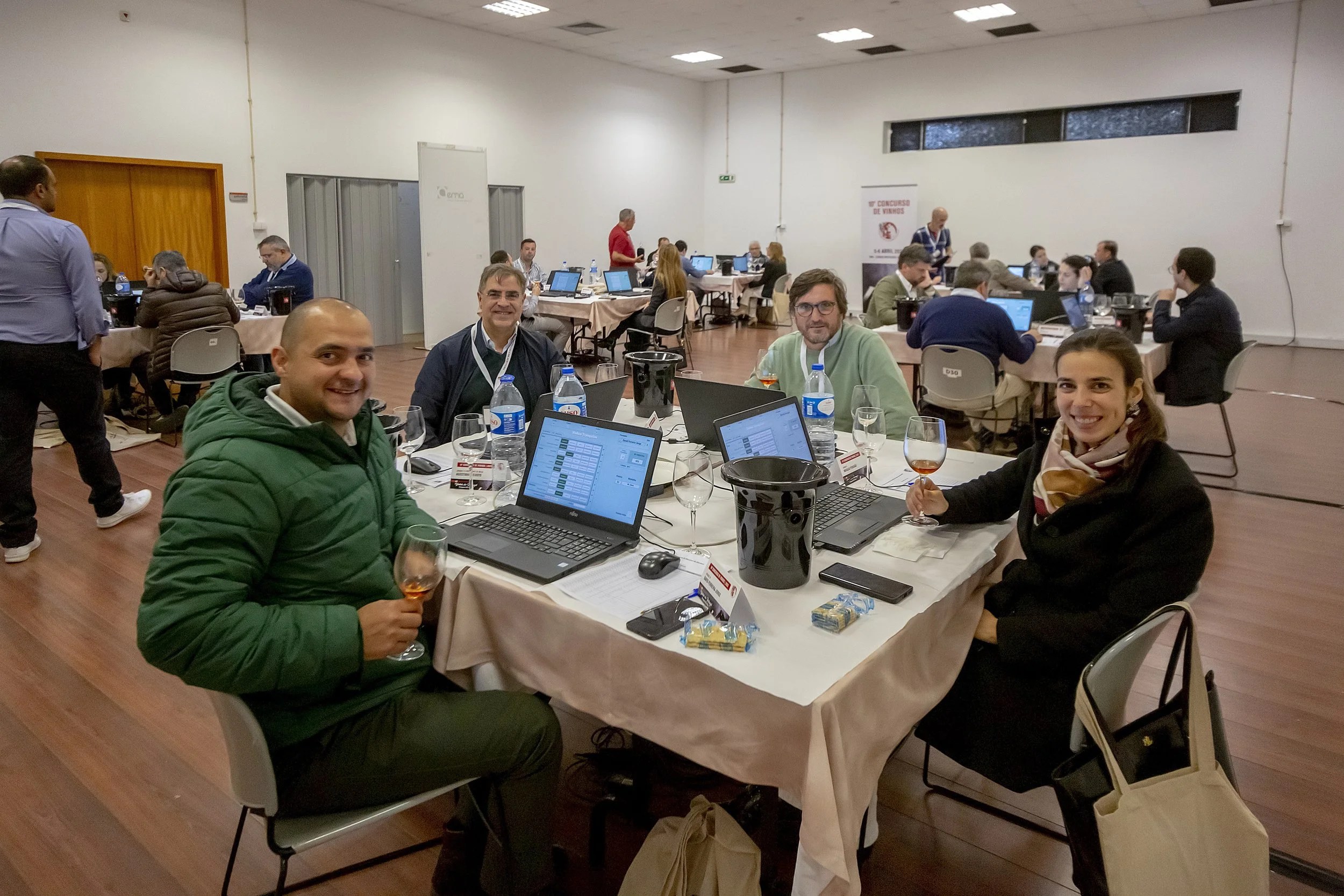 Grupo de pessoas sentado ao redor de uma mesa em uma conferência, com laptops, garrafas de água, taças de vinho e blocos de notas.