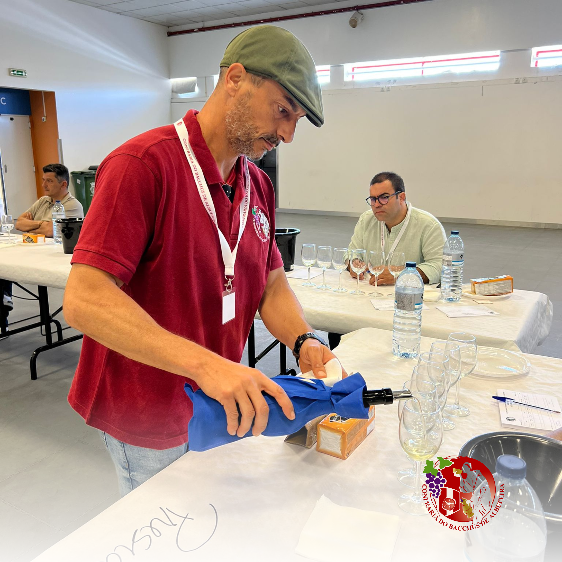 Homem abrindo uma garrafa de vinho em uma mesa de degustação com várias taças e garrafas de água. Outros participantes ao fundo observando.