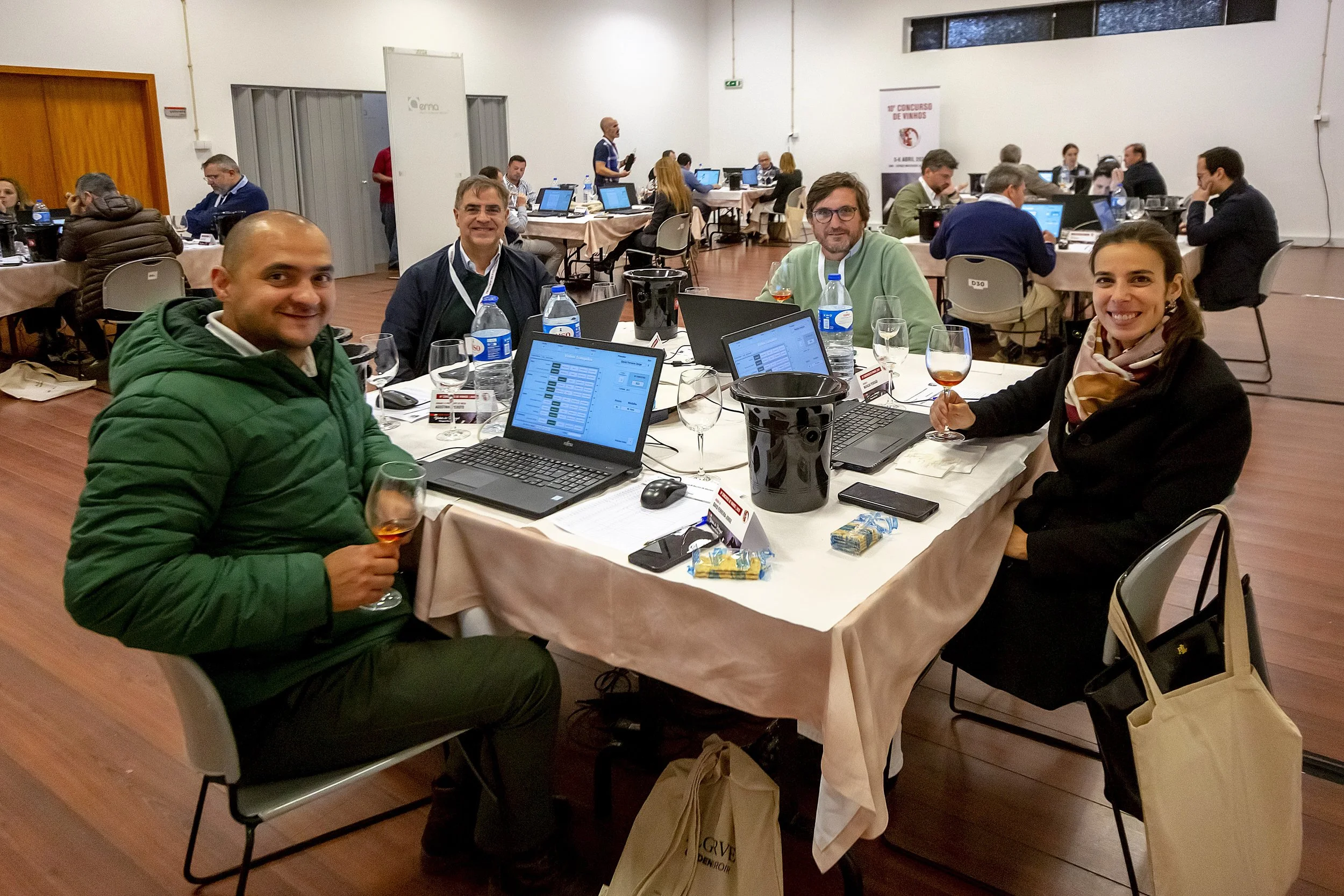 Grupo de pessoas participando de uma reunião ou conferência em uma sala de eventos, sentadas ao redor de uma mesa com laptops, bebidas e snacks.