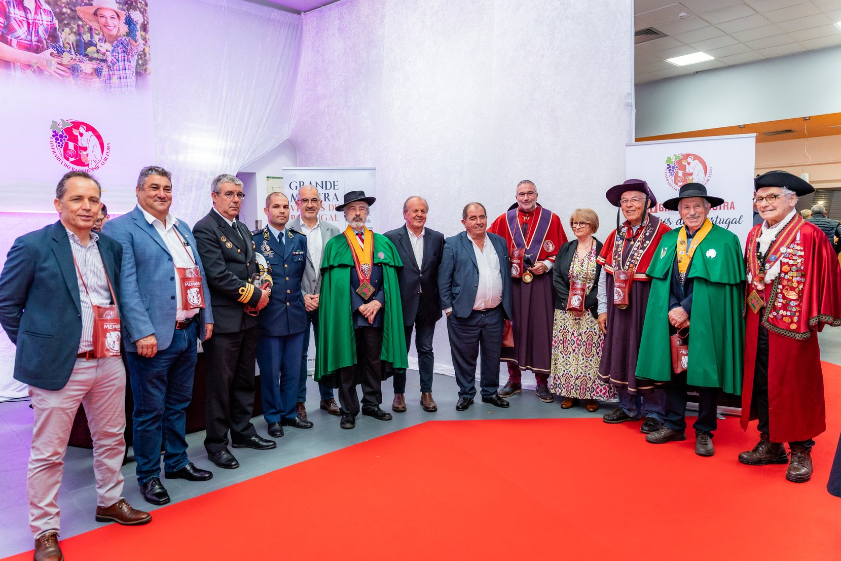 Grupo de pessoas, incluindo autoridades e membros de uma confraria, posando para uma foto de evento oficial em um ambiente interno com decoração em branco e painéis de fundo e uma faixa de tapete vermelho, alguns trajando roupas tradicionais e outras