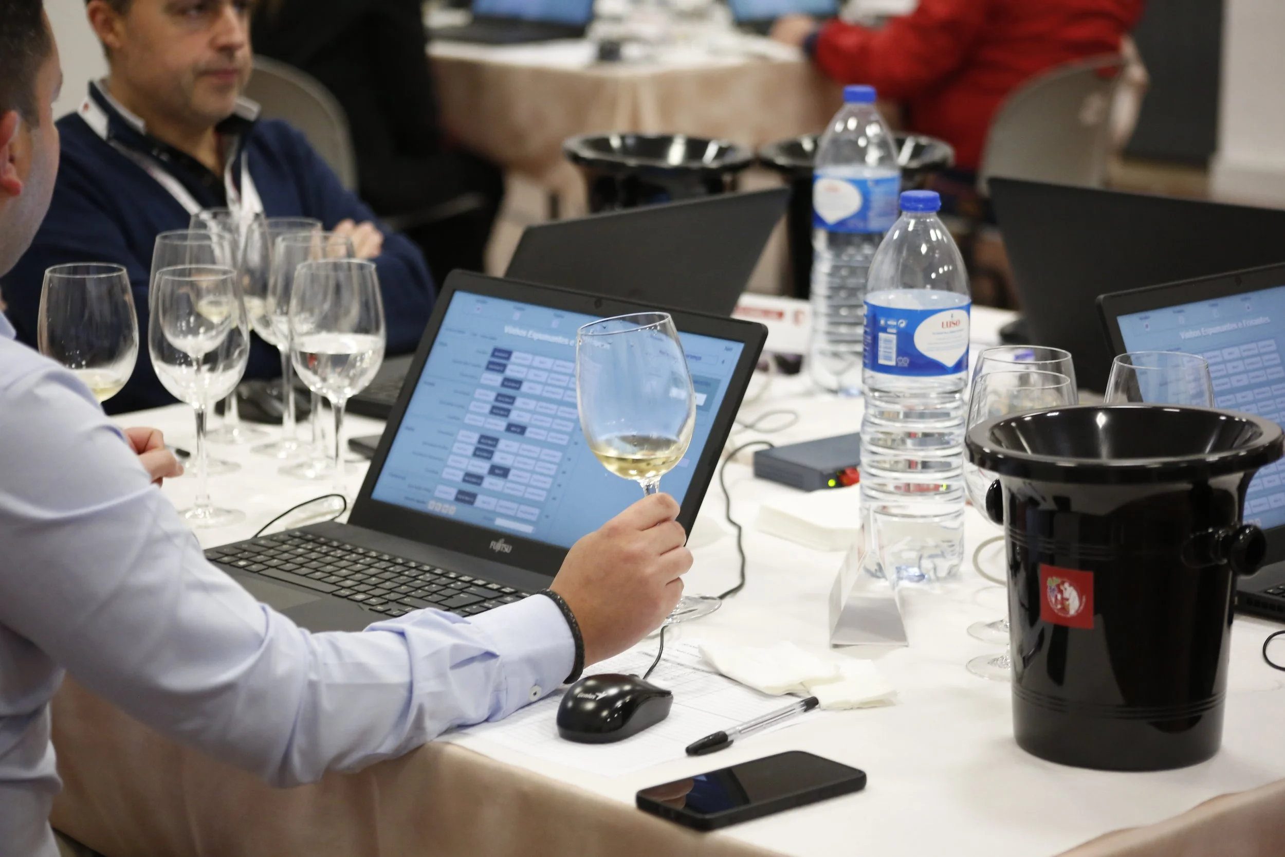 Reunião corporativa com várias pessoas usando laptops, garrafas de água e taças de vinho na mesa.