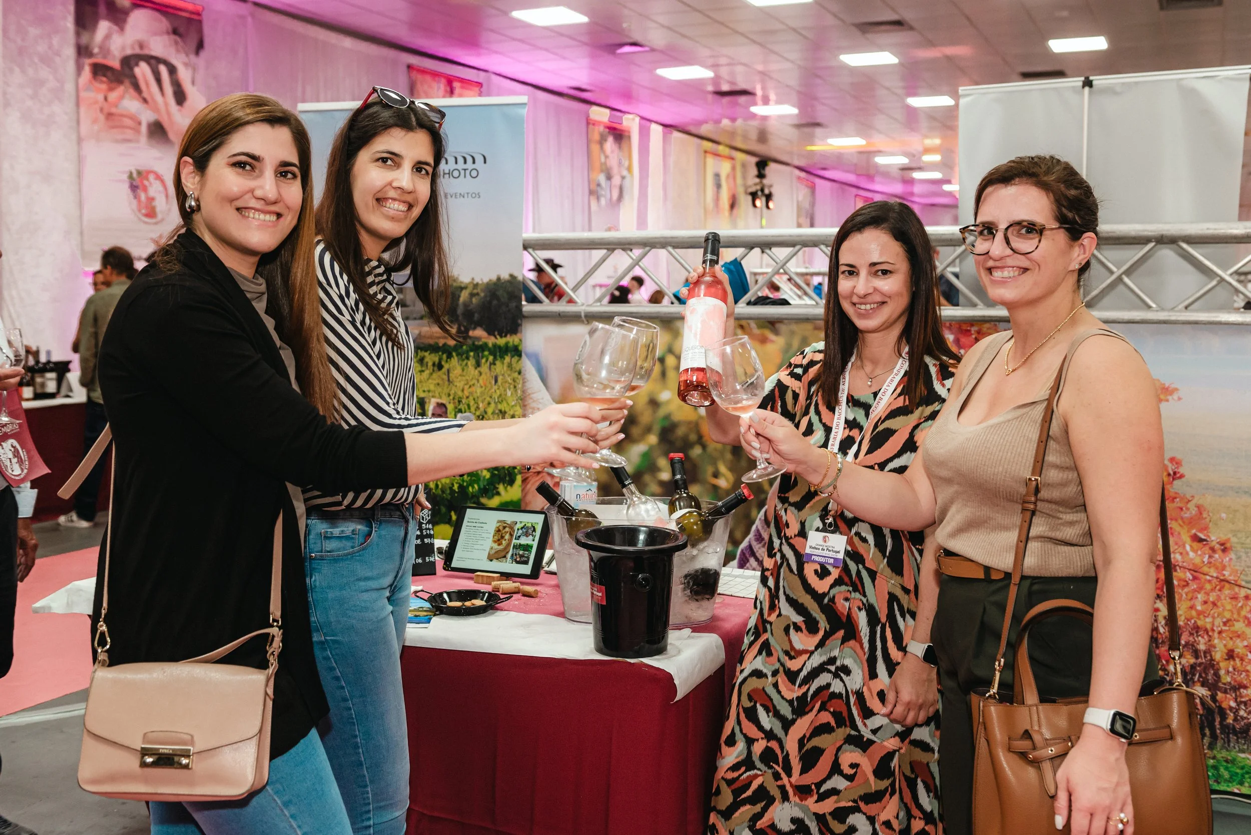 Quatro mulheres felizes brindando com taças de vinho em uma feira de vinhos, com uma mesa com garrafas e utensílios ao fundo.
