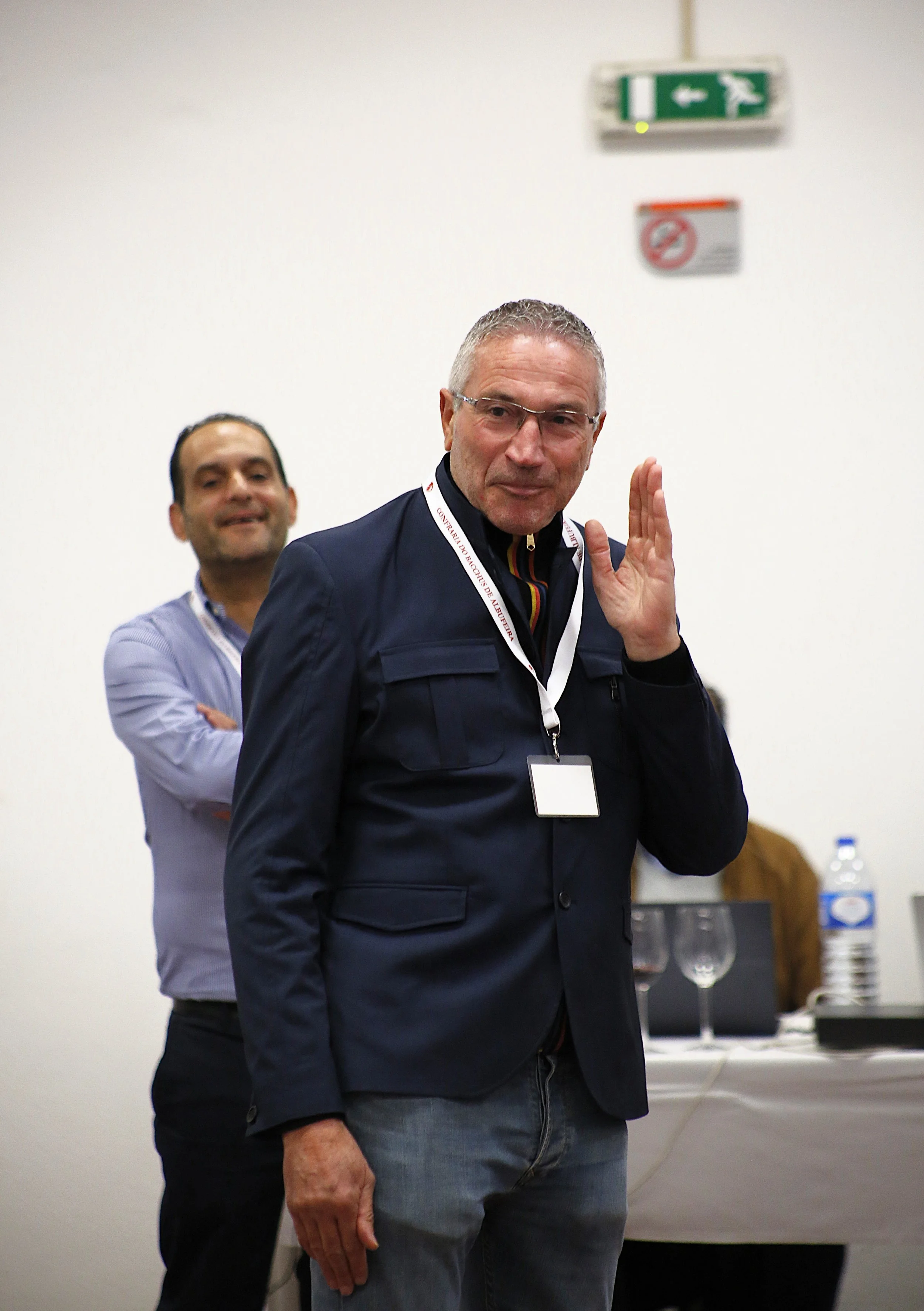 Homem com óculos de grau, cabelo grisalho, vestindo jaqueta azul e camiseta listrada, acenando, em ambiente de conferência ou reunião, com outro homem ao fundo ao lado de uma mesa com garrafas d'água e copos.