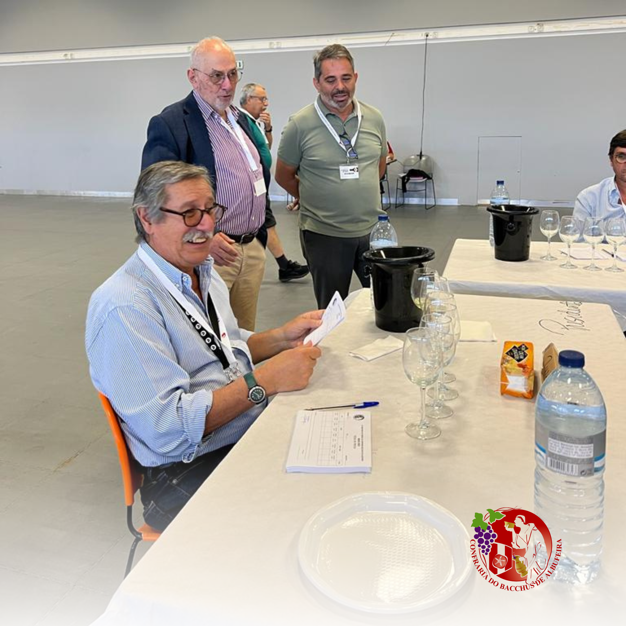Homens em uma reunião, um deles sentado na mesa segurando uma folha e sorrindo. Outros homens ao fundo observando, com taças de vinho e água, garrafas de água na mesa, e um logotipo de uma confeitaria na parte inferior direita da mesa.