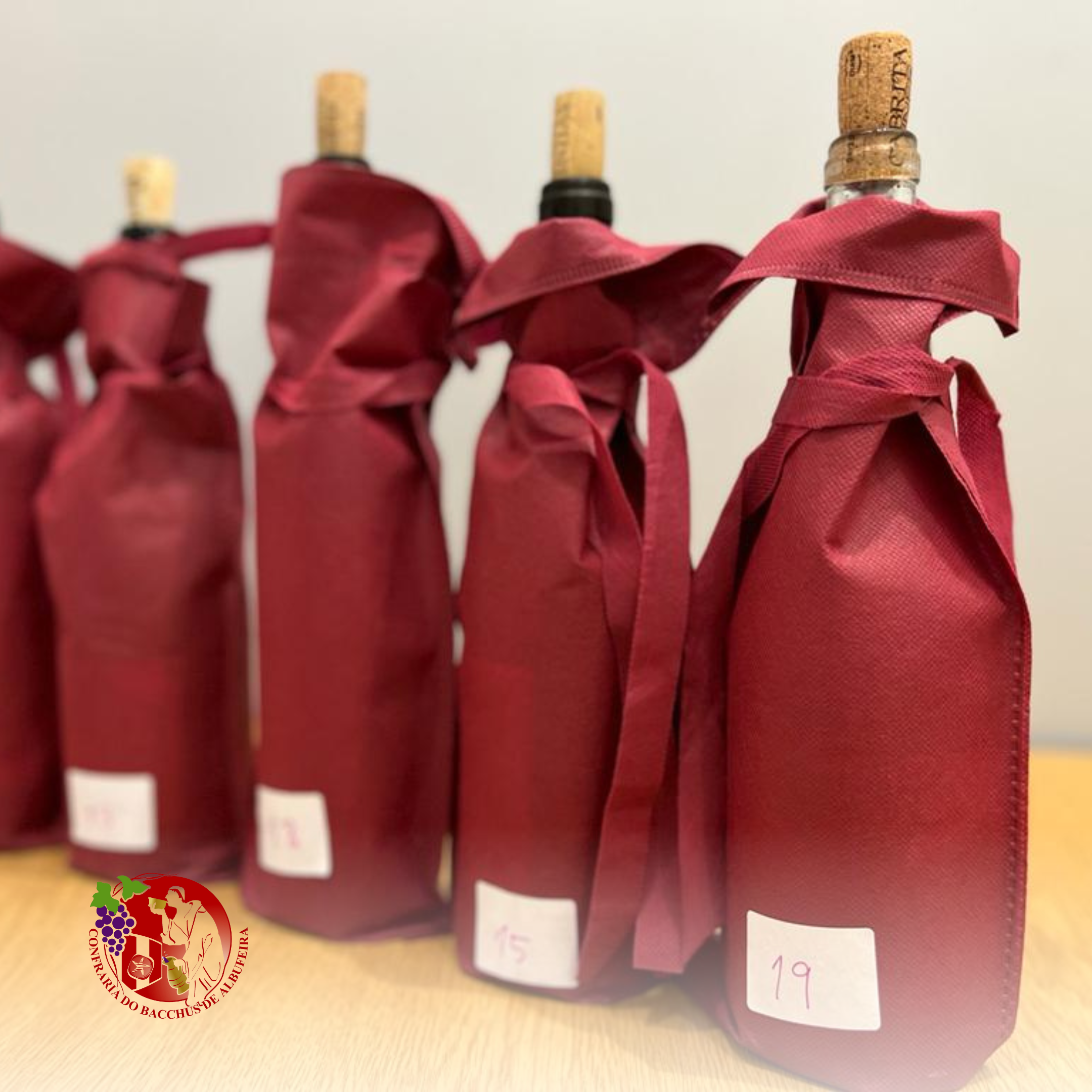 Garrafas de vinho cobertas com papel e fita vermelha, com etiquetas numeradas na frente, sobre uma mesa de madeira. Logo da Confraria do Bacchus de AlbuquerA.