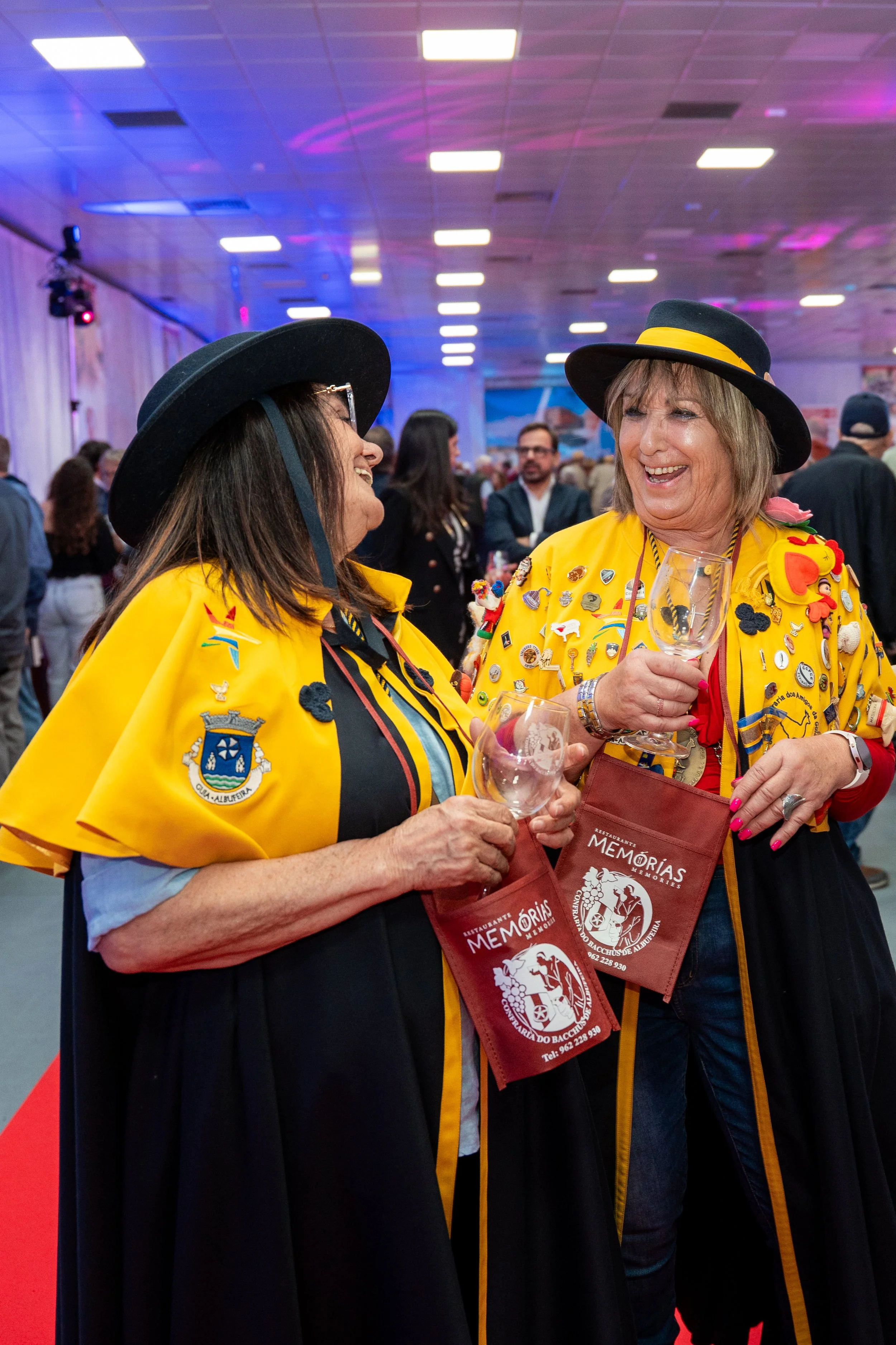 Duas mulheres sorrindo e conversando numa festa, usando roupas amarelas decoradas com patches e chapéus pretos, segurando taças de vinho.