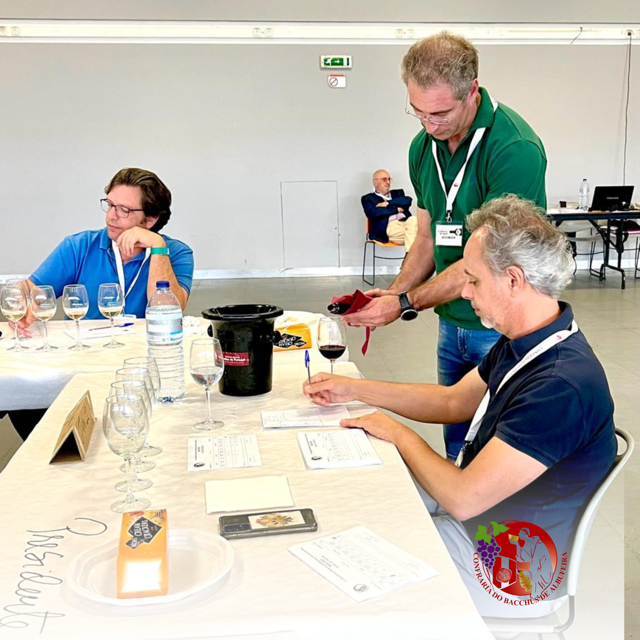 Três homens participando de uma degustação de vinhos, um deles escreve ou faz anotações. Há várias taças de vinho, uma garrafa de água, um balde para descarte de vinho, e uma tela de identificação na mesa. Um logo de evento de vinicultura está no can