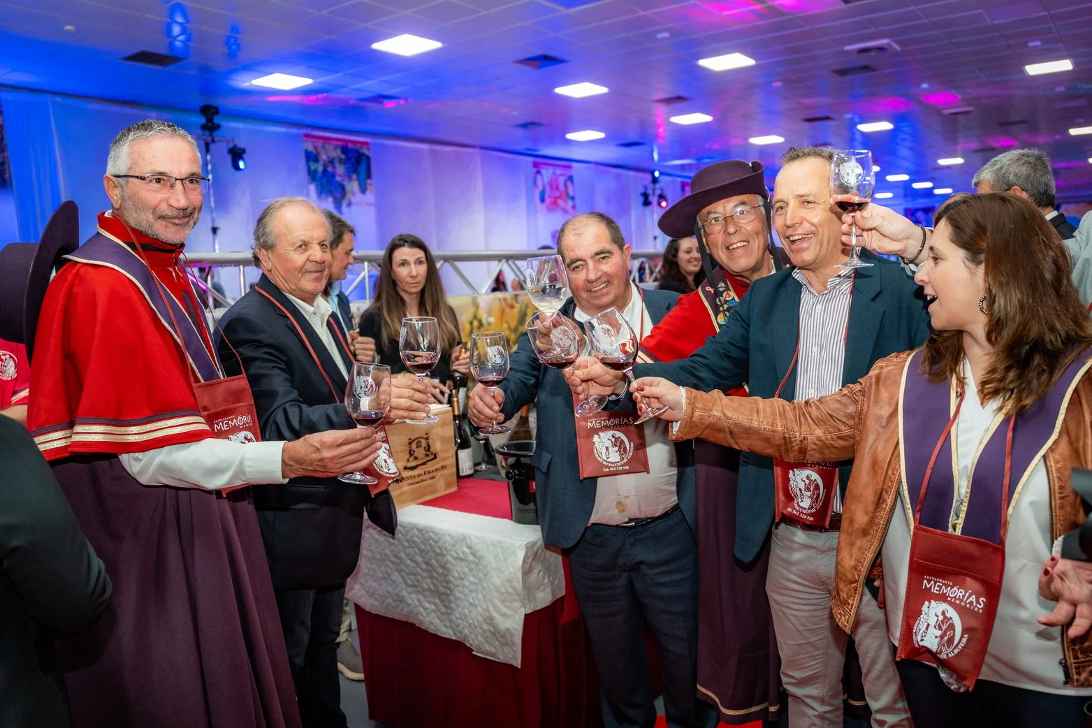 Grupo de pessoas celebrando com taças de vinho em evento social, alguns vestindo trajes tradicionais e outros mais casuais, ambiente com iluminação colorida e decoração festiva.