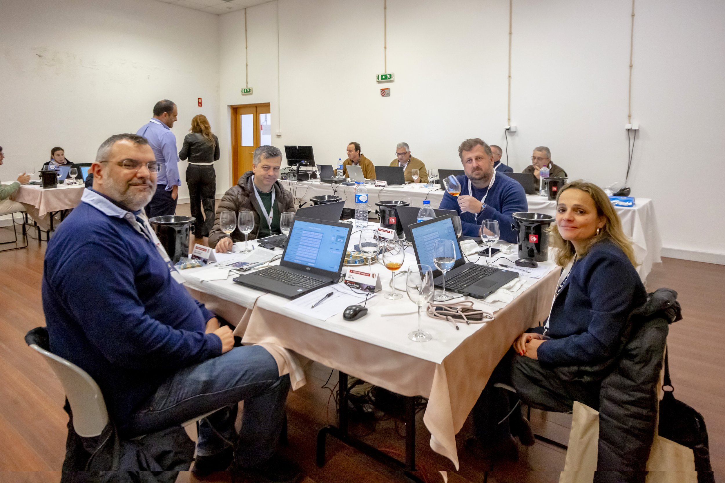 Reunião de várias pessoas ao redor de uma mesa com laptops, copos de vinho, garrafas de água, em um ambiente de conferência ou workshop