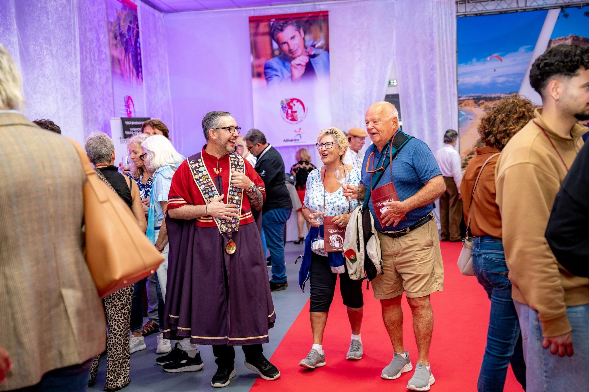 Pessoas conversando e sorrindo em uma feira ou evento cultural, com banners e fotos de fundo, incluindo uma grande foto de um homem, possivelmente um artista ou personalidade, em um ambiente bem iluminado e com uma decoração colorida.