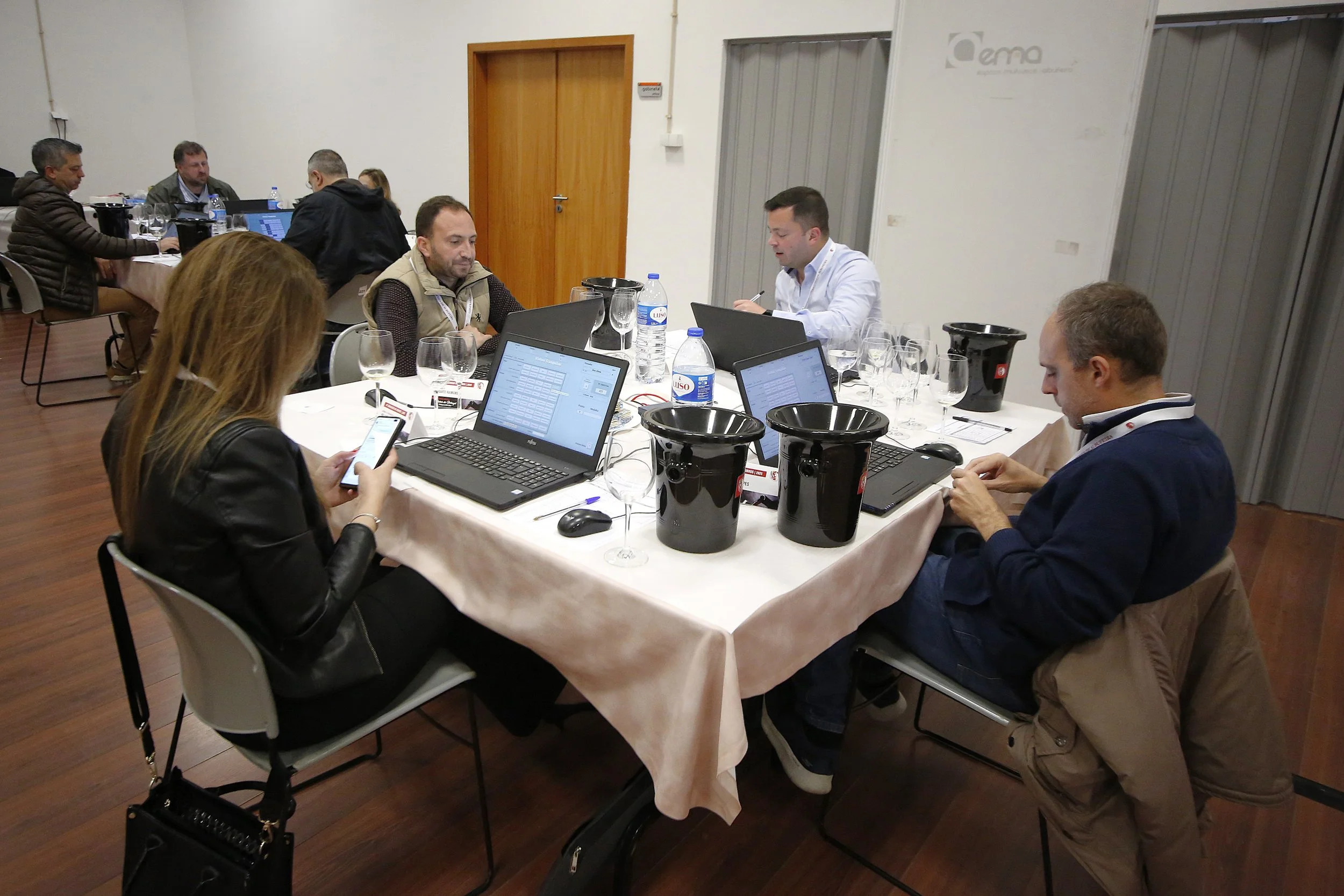 Reunião de pessoas em uma sala de conferência, sentadas ao redor de uma mesa com laptops, garrafas de água e copos, participando de um evento de trabalho ou treinamento.