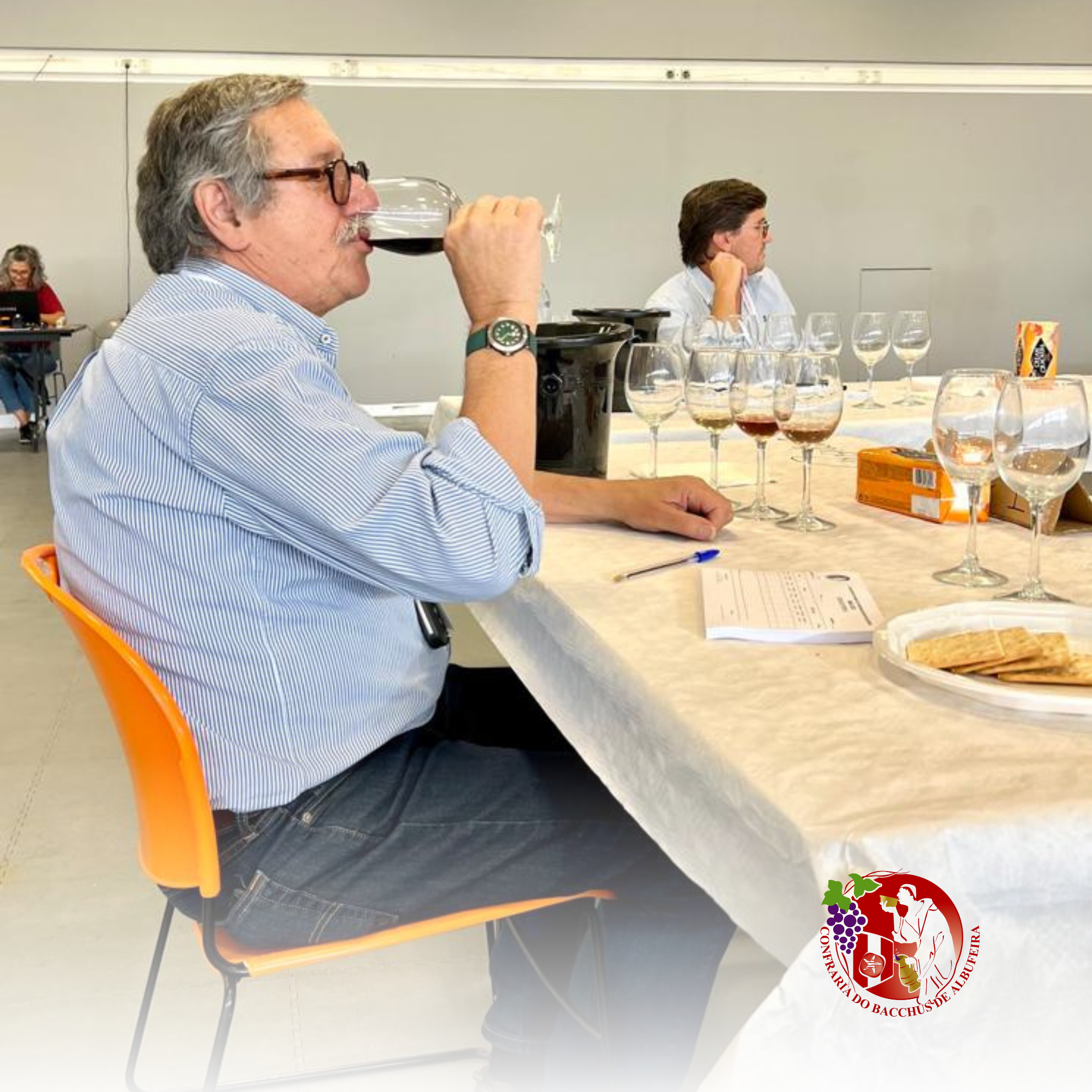 Homem de cabelos grisalhos, óculos, camisa de listras, sentado em uma mesa de degustação de vinhos, segurando uma taça de vinho tinto e cheirando o conteúdo, com várias taças de vinho em frente, logo da Confraria do Bacchus de Albufeira no canto infe