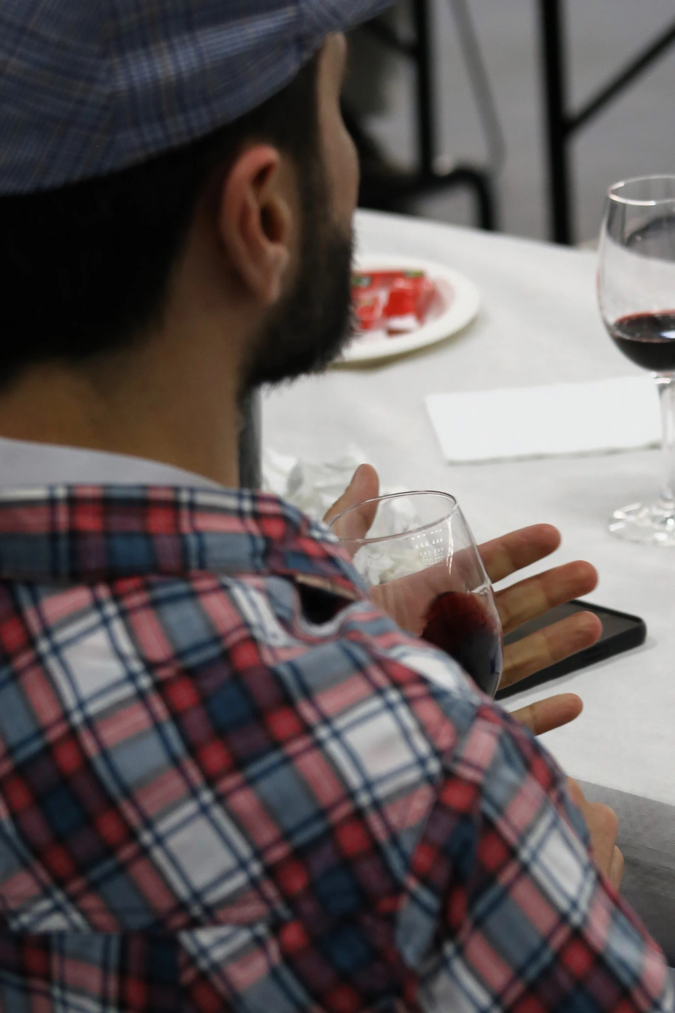 Homem de camisa xadrez vermelho e azul segurando uma taça de vinho tinto em uma mesa com copos de vinho e pratos ao fundo.