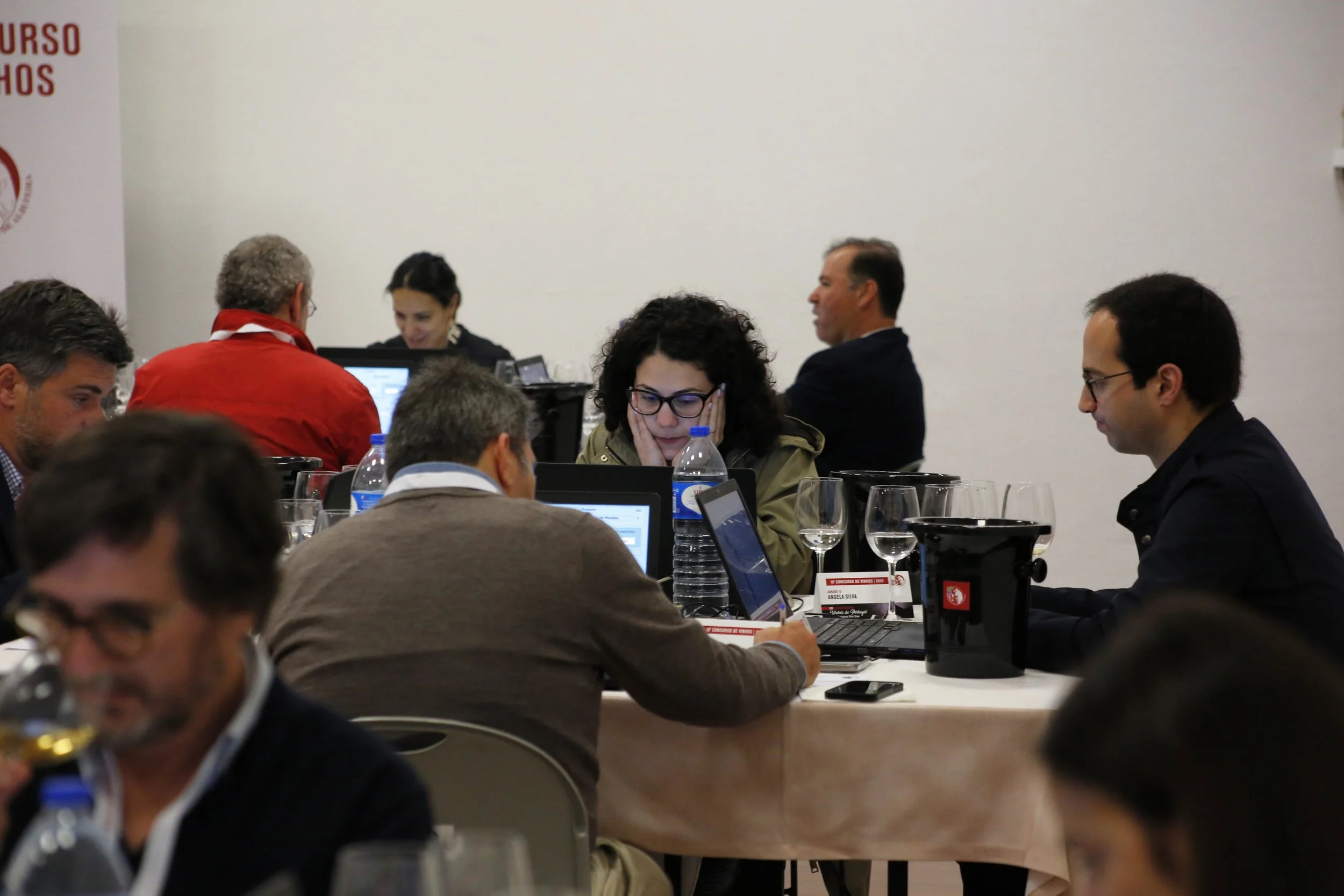 Pessoas sentadas ao redor de uma mesa de reunião, usando laptops e garrafas de água, em um ambiente de conferência ou seminário.
