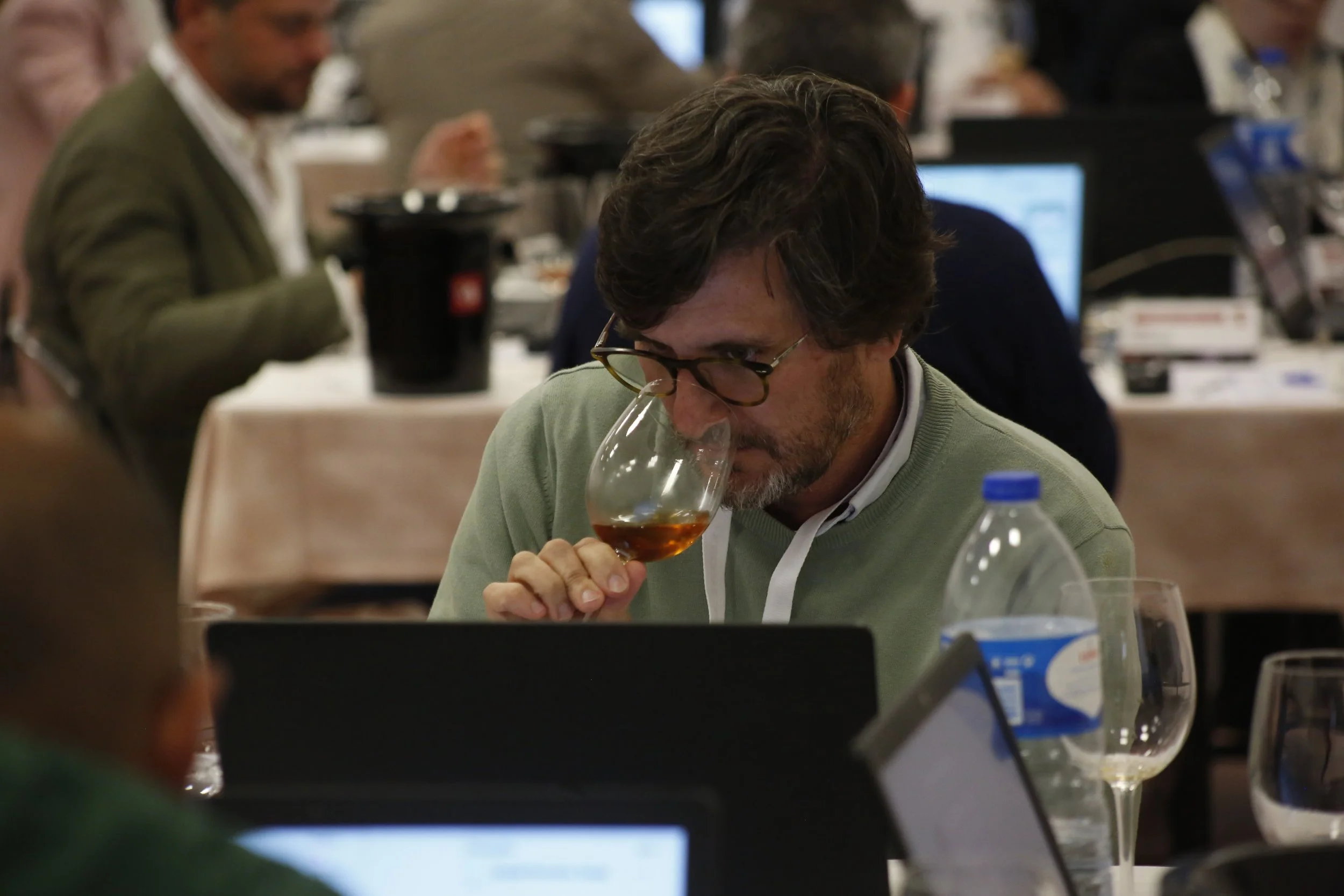 Homem com óculos de armação escura, barba e cabelo castanho claro, provando uma taça de vinho tinto em uma conferência ou evento. Ao redor, há várias pessoas concentradas, utilizando laptops, em um ambiente de sala de reuniões.