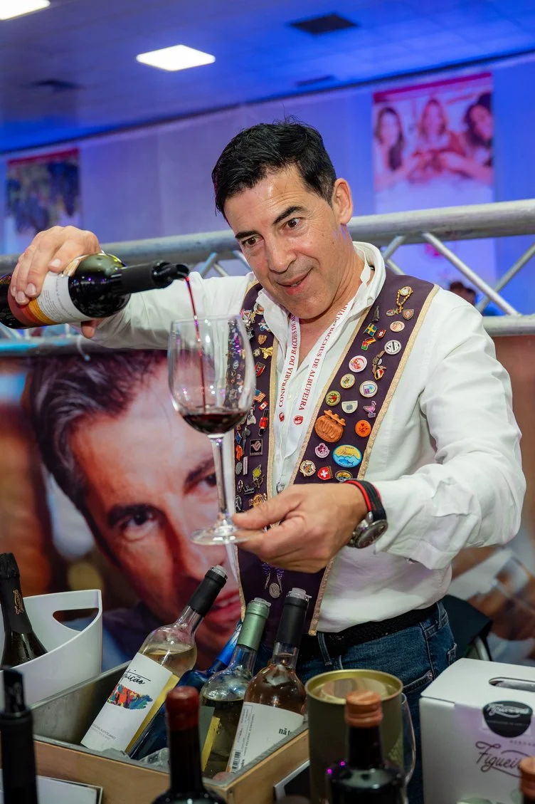 Homem servindo vinho tinto em taça e vestindo uniforme decorado com muitos bottons, ao fundo uma parede com pôster de uma pessoa e uma mulher.